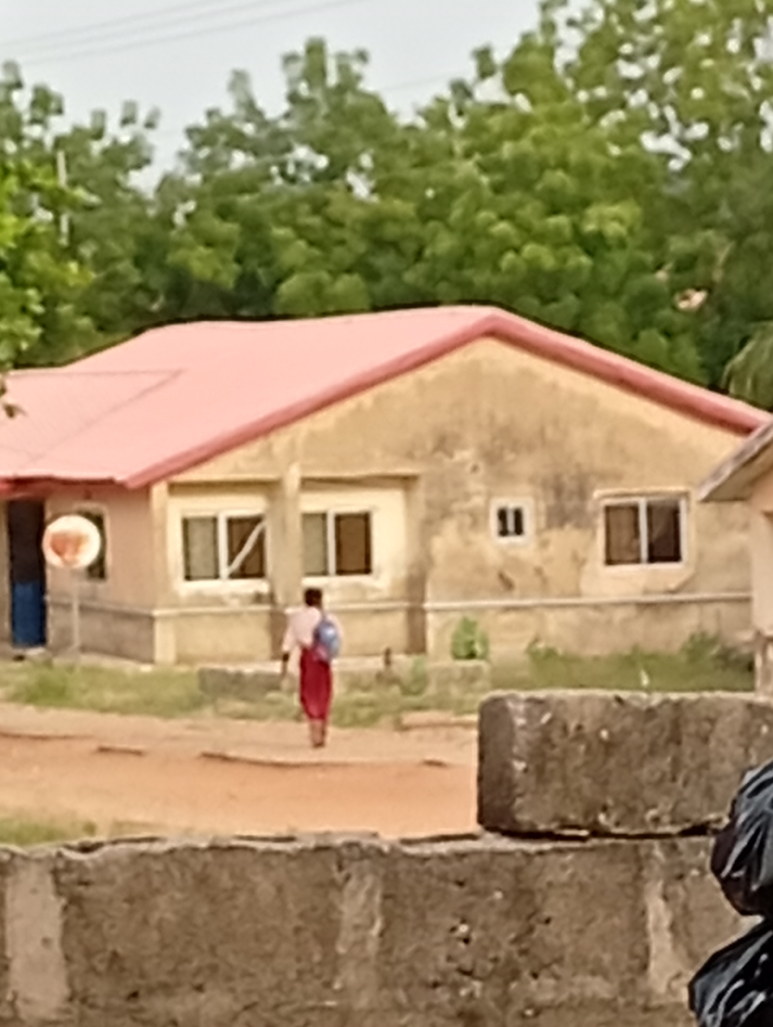 A residential area is visible in Unknown, Unknown. A building with a light beige exterior and a salmon-colored roof is in the background. The building has multiple windows with dark interiors. A basketball hoop is attached to the side of the building near a blue door. A person wearing a light-colored shirt and a red skirt or pants walks on a dirt path in front of the building, carrying a blue bag. In the foreground, there is a low wall made of rough, gray blocks. Green trees are in the background behind the building. The sky is overcast.
