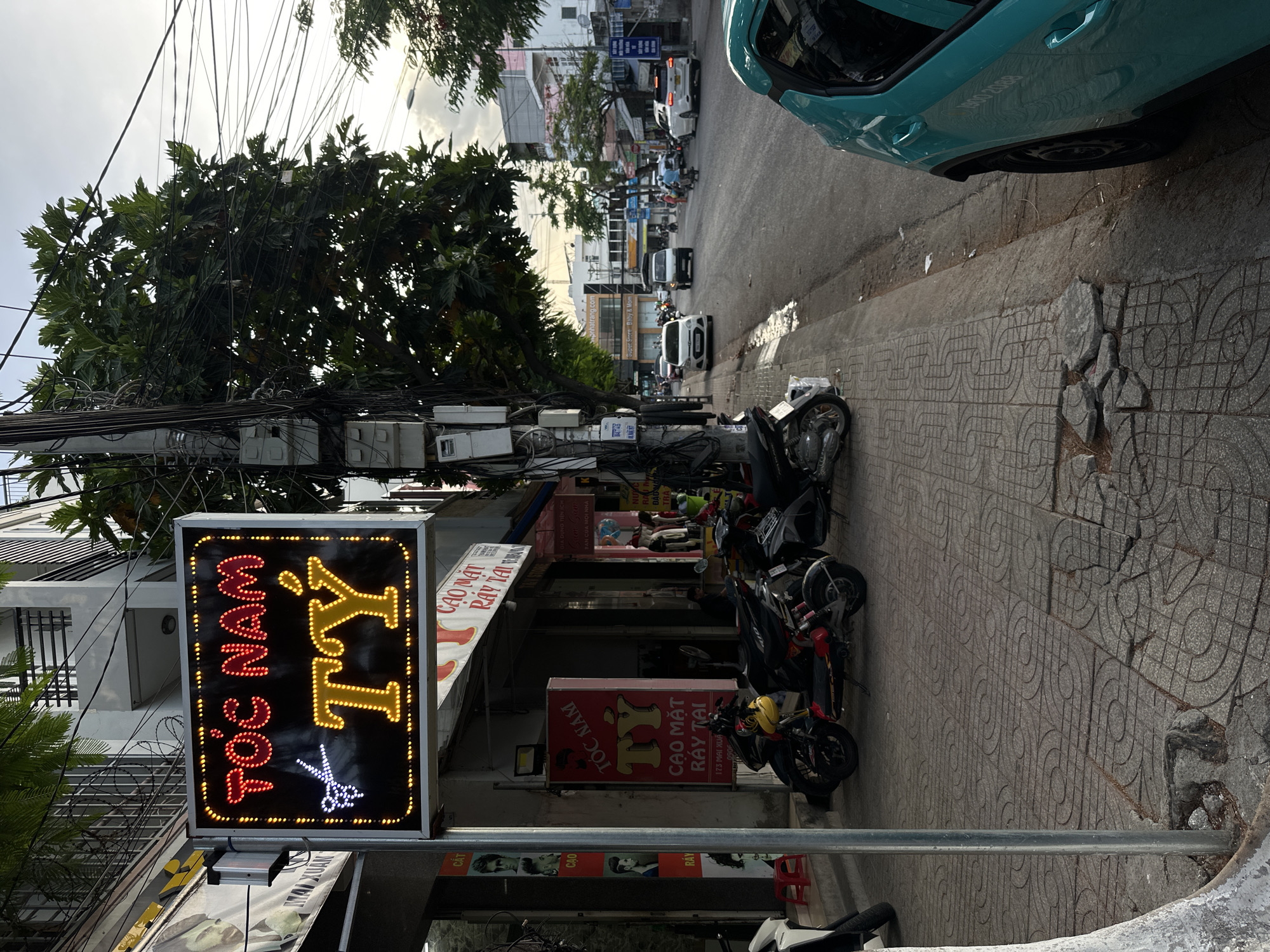 The image depicts a street scene in Bac Nha Trang Ward, Vietnam. In the foreground, a neon sign reads "TÓC NAM TÝ" in red and yellow, featuring a pair of scissors in blue and white. The sign is mounted on a metal pole. To the left of the sign is a sidewalk paved with patterned tiles, with some broken sections. A white motorcycle is partially visible on the far left. Several other motorcycles are parked along the sidewalk. A tall pole with numerous electrical wires and a few utility boxes stands to the right of the sign.
The street itself is asphalted and appears to be moderately busy with vehicular traffic, including cars and motorcycles. The buildings lining the street are of varied architectural styles, with some featuring balconies and awnings. Lush green trees with dense foliage are visible, casting shadows on the sidewalk and street. The sky is overcast, suggesting it may be late afternoon or early morning. The overall impression is of a typical urban street in Vietnam, characterized by its bustling atmosphere, utility infrastructure, and the presence of motorbikes.