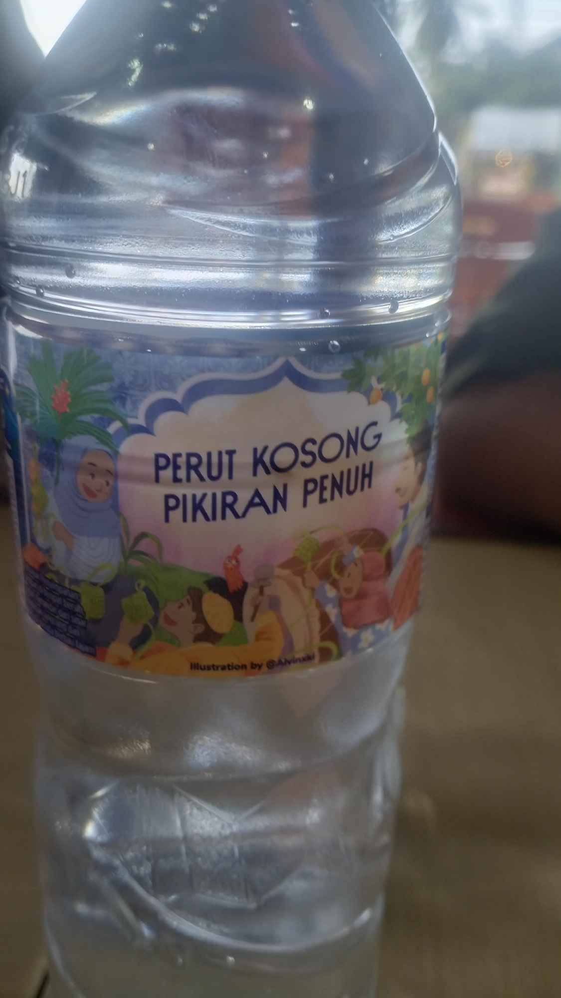 A close-up shot captures a clear plastic water bottle, likely containing purified water, in what appears to be a casual indoor or sheltered outdoor setting in Lhokseumawe, Indonesia. Condensation and water droplets are visible on the exterior of the bottle, suggesting a cool temperature inside relative to the ambient environment.
The bottle features a decorative label with cartoon-like illustrations set against a light blue patterned background. The central part of the label, shaped like a cloud, prominently displays text in Indonesian: "PERUT KOSONG PIKIRAN PENUH," which translates to "Empty stomach, full mind." Below this text, in a smaller font, is "Illustration by @Alvinxxi," crediting the artist.
The illustrations depict several people, rendered in a cheerful, whimsical style. On the left, a woman wearing a light blue hijab is shown, smiling. Various other characters, including children and adults, are integrated into a lush green landscape featuring foliage and trees bearing orange fruits. Some characters appear to be lying down, looking up, or interacting with the natural elements. The scene conveys a sense of calm and contentment. The blurred background shows indistinct shapes that could be furniture or parts of a building.