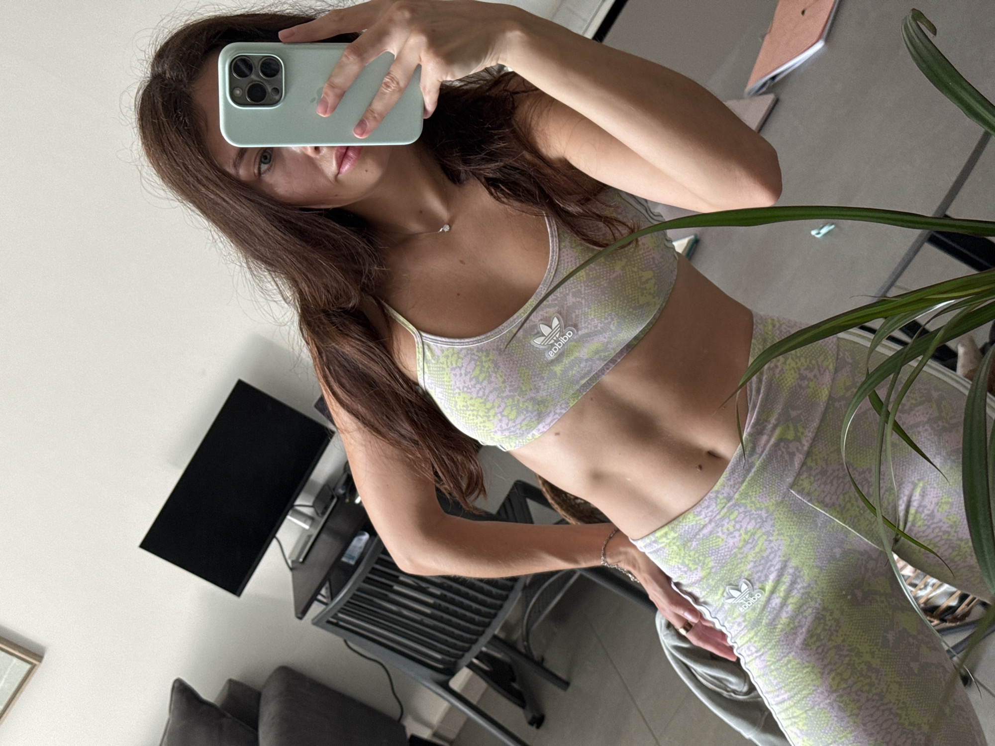 The image shows a young woman taking a selfie in front of a mirror. She is wearing a matching Adidas sports bra and leggings with a pale green and lilac snakeskin print. Her long brown hair falls over her shoulder. She is holding a light green smartphone with multiple camera lenses in front of her face, obscuring part of her face.
The background reveals a modern interior. A black television is mounted on the wall above a dark grey metal desk or table. Below this, there is a grey couch or seating area. A potted plant is partially visible in the foreground on the right side of the frame. The location cannot be determined.