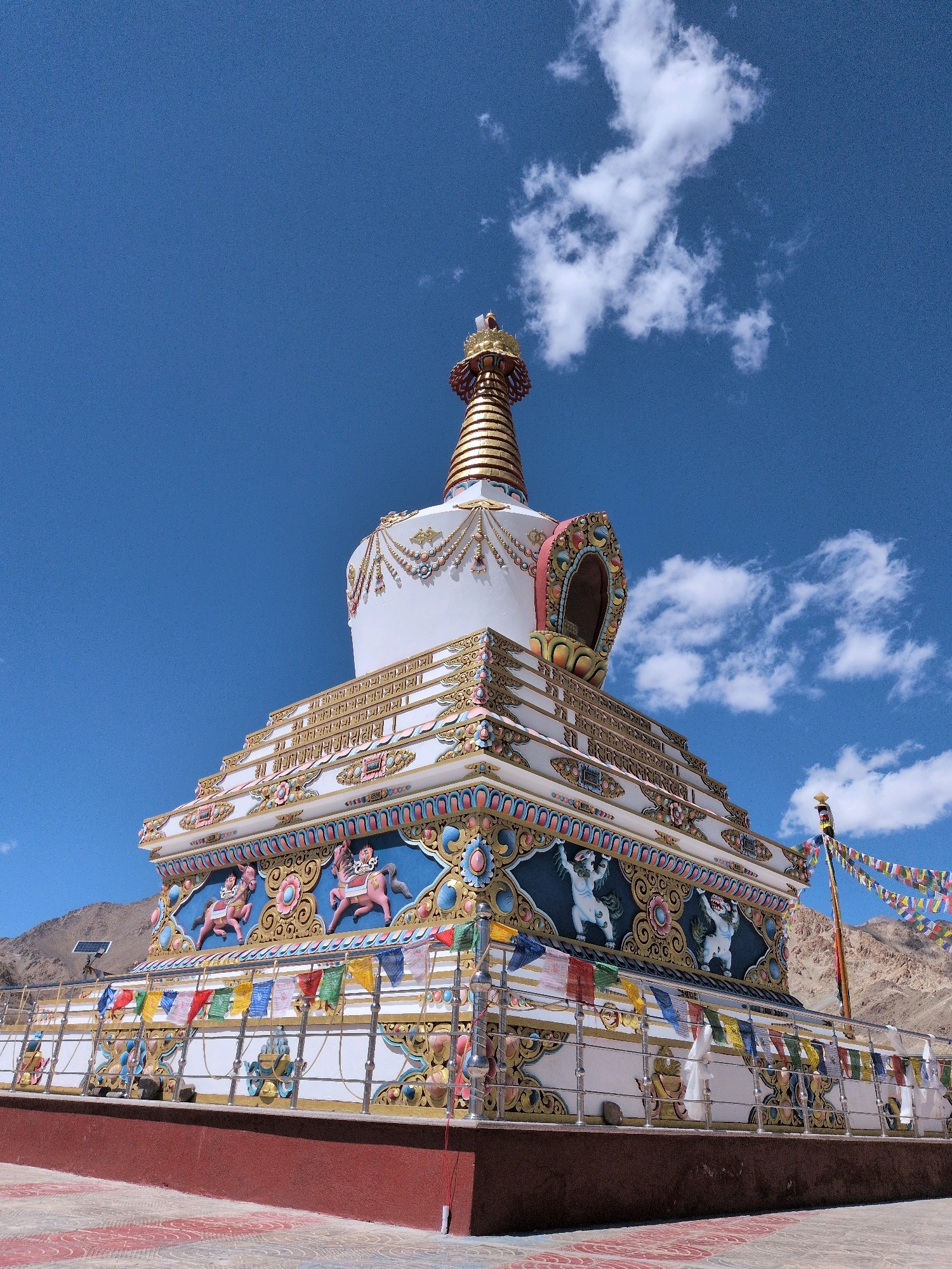 A Buddhist stupa, painted white with ornate gold and colorful accents, stands against a clear blue sky with scattered white clouds. The stupa features intricate carvings and decorations, including images of horses and mythical creatures. Prayer flags in various colors flutter from a pole on the right side of the structure. The stupa is situated on a raised platform with a red base and a patterned tiled ground. In the background, dry, rocky hills are visible. This is the Shanti Stupa in Leh, India.