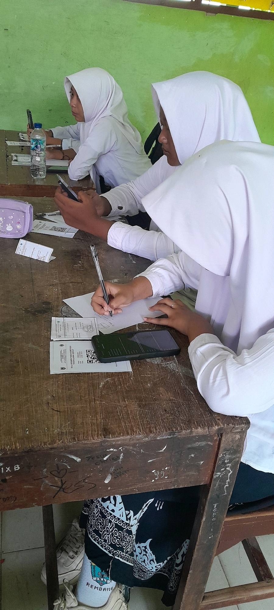 The image shows two female students wearing white hijabs and white shirts, seated at a wooden desk in a classroom. The student in the foreground on the right is writing on a piece of paper with a pen, while holding a smartphone in her left hand. In front of her are several pieces of paper, one with a QR code, and a smartphone. The student on the left is looking at her smartphone, with a bottle of water and another smartphone on the desk in front of her. The classroom environment appears simple, with a green wall in the background. The desk is old and worn, with graffiti visible underneath. The students are likely in a school in Tahunan, Indonesia, given the context provided.