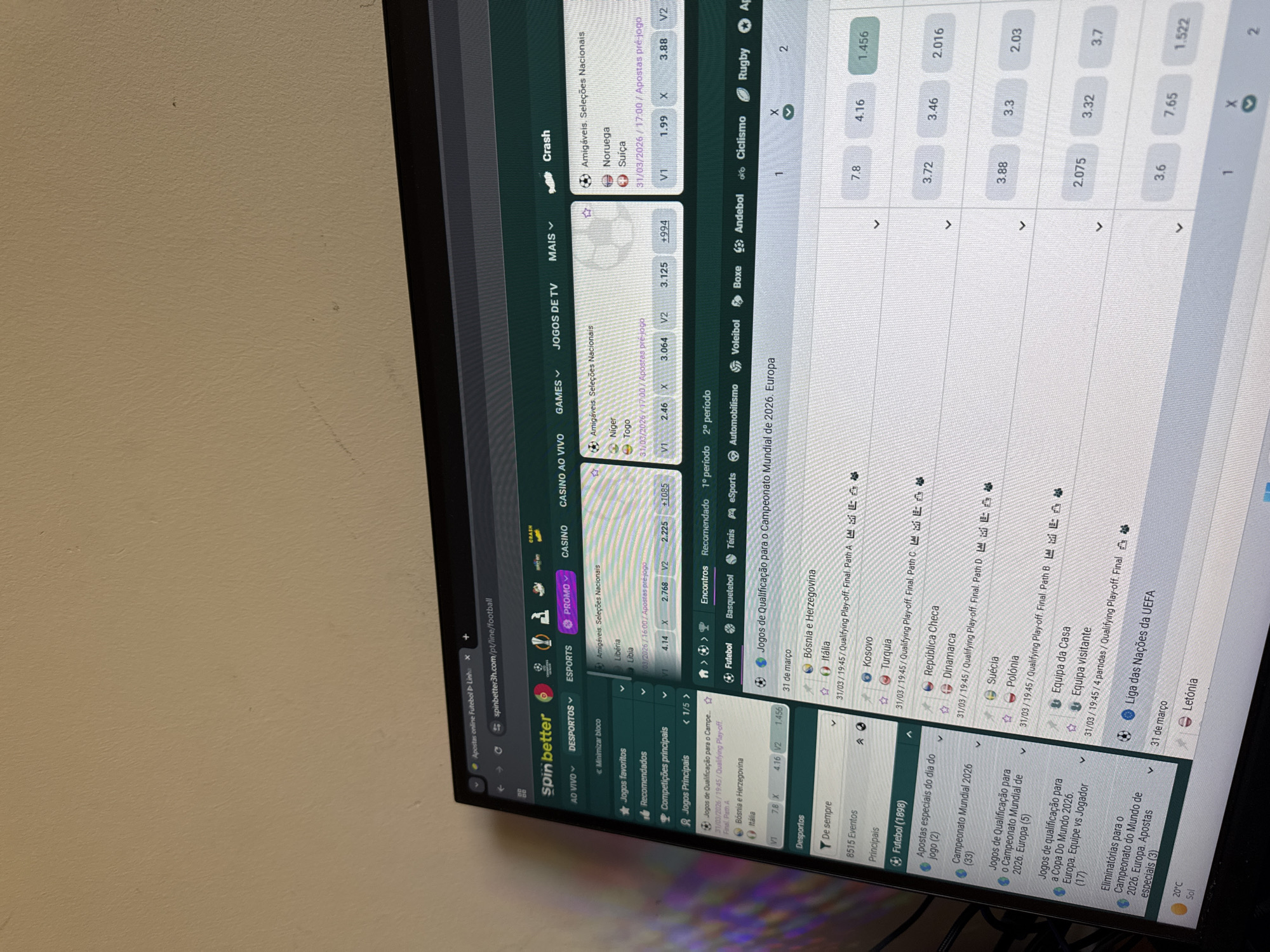 The image depicts a computer screen displaying a betting website called "SpinBetter". The website features various sports games and odds, including soccer, tennis, cycling, and rugby. The games include qualifying matches for the World Cup 2026, the UEFA Nations League, and friendly matches. On the left, menus and options related to sports and casino games are visible. The computer screen is positioned in front of a beige wall.