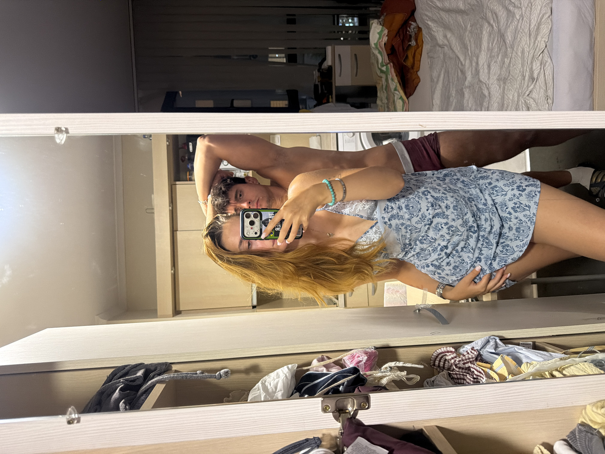 A young woman with long, blonde hair is taking a mirror selfie in a bedroom. She is wearing a light blue, floral print dress and holding a phone in her right hand. A young man with short, dark hair is standing behind her, with his left arm raised behind his head and his right arm resting on his hip. He is wearing maroon underwear. The room appears to be a shared living space, with a closet filled with clothes on the left and a made bed visible in the mirror on the right. The overall impression is of a casual, intimate moment captured in a domestic setting. This image was taken in Sofia, Bulgaria.