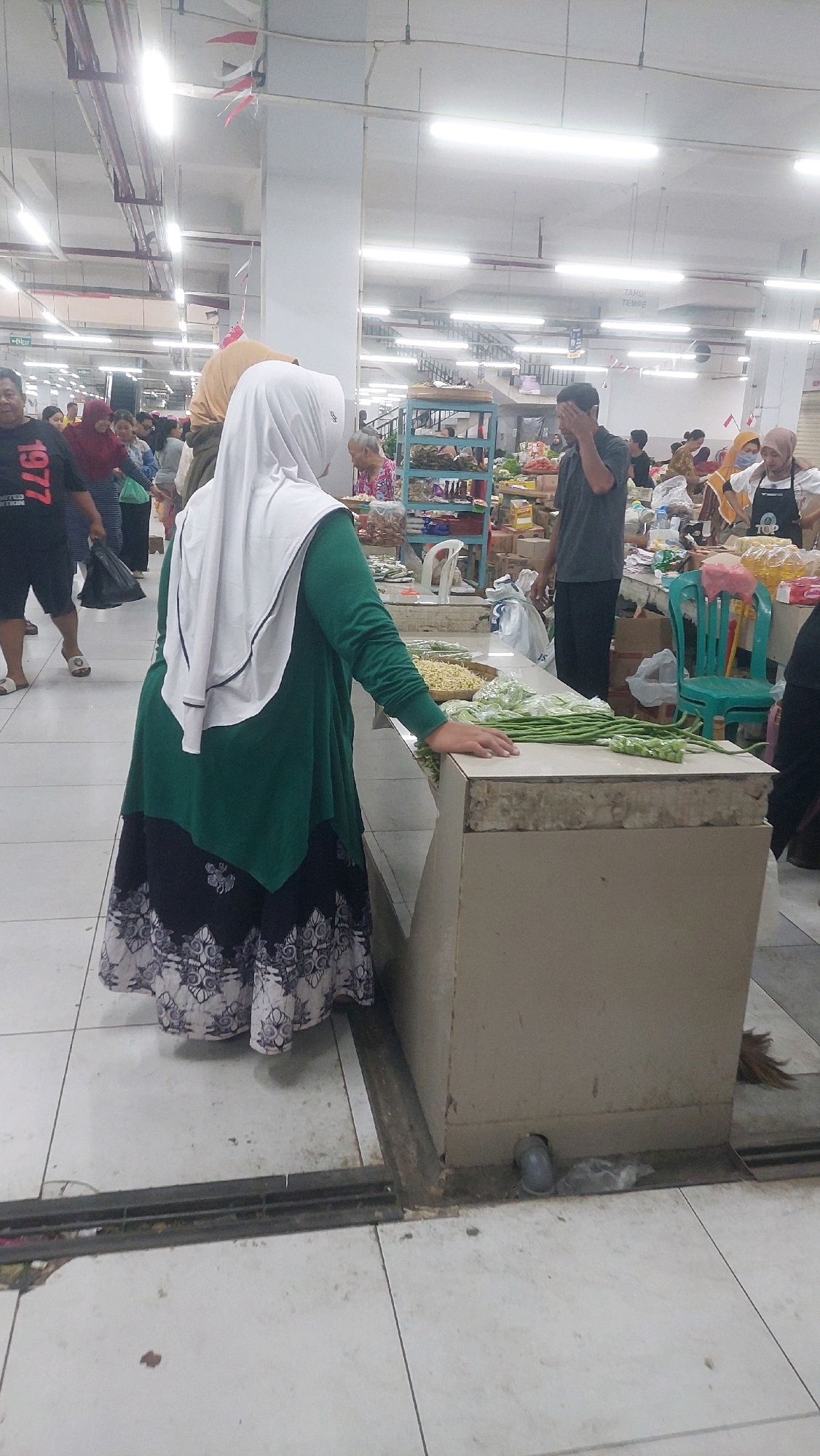 The image shows a bustling indoor market scene. In the foreground, a woman wearing a green dress and a white hijab is standing at a market stall. Her hand is resting on a counter where piles of fresh green vegetables, possibly long beans, and a bowl of dried noodles are displayed. The stall itself is a large, beige structure with a worn edge. The flooring is tiled.
In the background, the market is filled with people, some of whom are obscured by the foreground elements. Market stalls with various goods, shelves, and lighting fixtures are visible. Signs with Indonesian text are present on some stalls, such as "TAHU TEMPE" (Tofu Tempeh) on a blue shelf unit. The overall atmosphere suggests a busy, local marketplace. The context of Ponorogo, Indonesia, indicates this is likely a traditional wet market or a supermarket with a fresh produce section.
