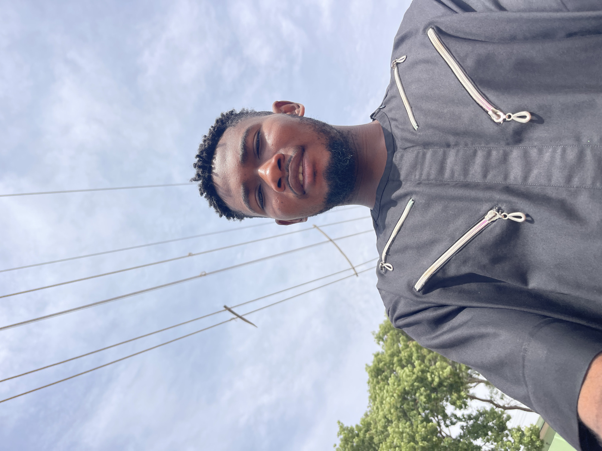 A low-angle, portrait-style photograph, rotated 90 degrees clockwise, captures a young Black man outdoors. He has short, dark, textured hair (resembling locs or dreadlocks), a well-kept beard, and is looking directly at the camera with a calm, pleasant smile.
He is dressed in a dark-colored, long-sleeved garment, likely a traditional tunic or kaftan, which features several decorative zippers on the chest and shoulder. These zippers have light-colored (cream or white) tapes and metallic silver-toned pulls with a looped design. A subtle pink hue is visible on the tape of one zipper.
The background predominantly features a bright, clear blue sky with scattered white clouds, indicating daytime. Several thin power or telephone lines crisscross the sky horizontally behind the man. In the lower right section of the image, a cluster of green trees with dense foliage is visible. The scene suggests an outdoor setting in Muchiya, Nigeria. No visible text is present in the image.
