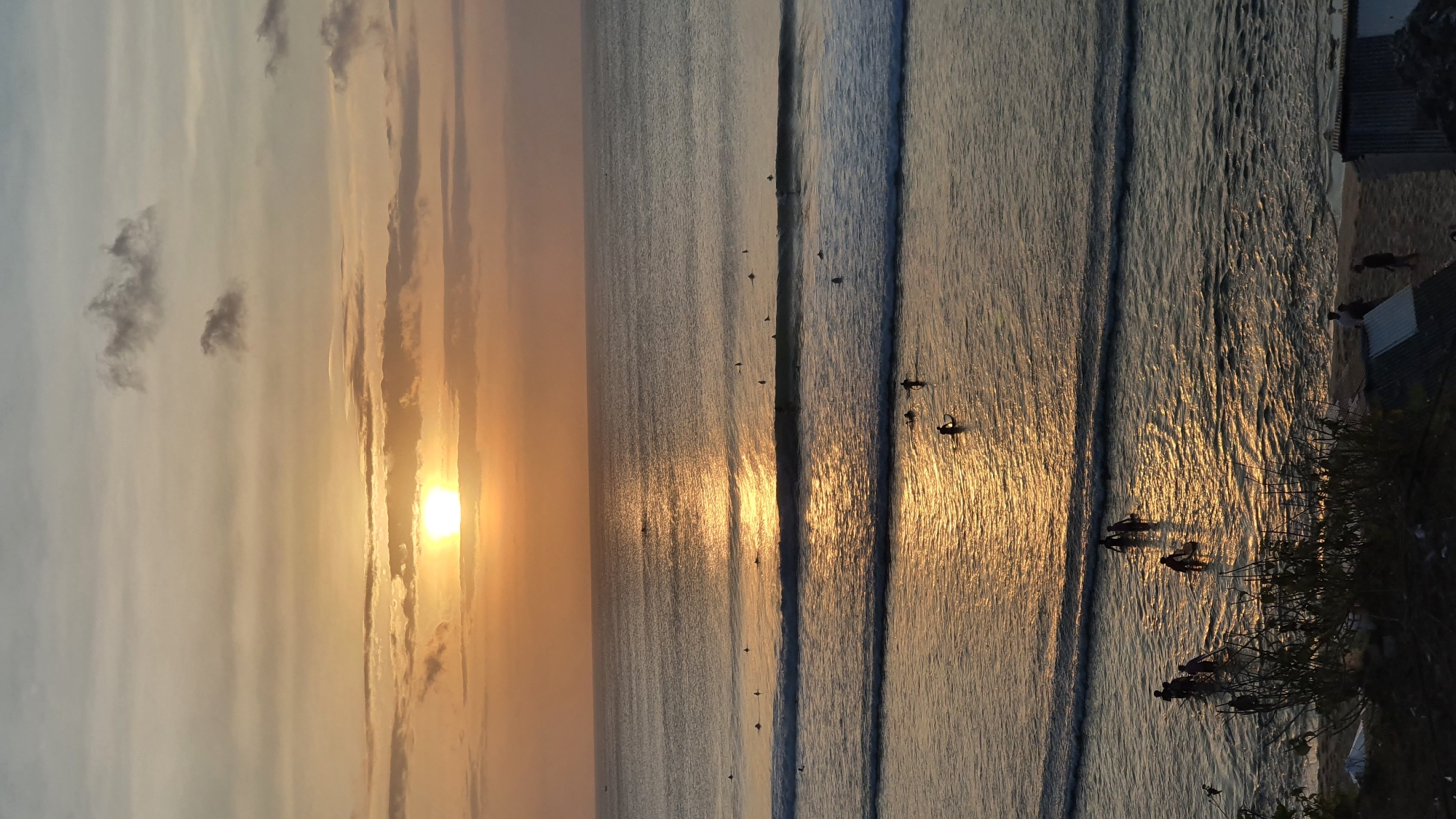 The image depicts a coastal scene at sunset in Kuta Selatan, Indonesia. The sun, low on the horizon, casts a warm, golden glow across the ocean, illuminating the gentle waves. Wispy clouds streak across the sky, painted in hues of orange and pink. A group of surfers are scattered across the water, their silhouettes visible against the shimmering surface as they wait for or ride the waves. Some are standing on their surfboards, while others are paddling. On the shoreline, a few people can be seen walking along the beach, their figures small against the vastness of the ocean. Buildings and vegetation are visible on the far right of the image, suggesting the proximity of human habitation to the beach. The overall mood is serene and peaceful, capturing the tranquil beauty of a tropical sunset. There is no visible text in the image.