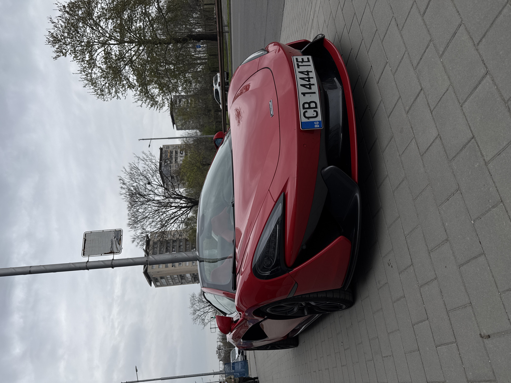 The image shows a red McLaren sports car parked on a gray brick sidewalk. The car's license plate reads "CB 1444 TE" with the "BG" country code indicating Bulgaria. The car is parked in what appears to be a city setting. Behind the car, there are trees, buildings, and a street sign on a pole. Based on the license plate and the general architectural style, the location is likely somewhere in Bulgaria, possibly in the city of Plovdiv.