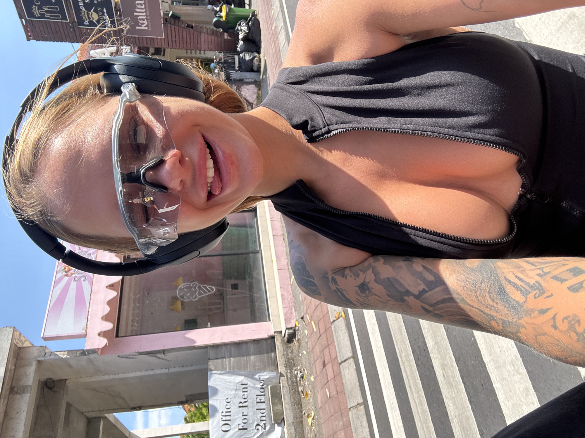 A woman is taking a selfie while standing on a crosswalk in North Kuta, Indonesia. She is wearing black headphones and transparent safety glasses. She has a black zippered top, with the zipper partially undone. Her left arm is heavily tattooed. In the background is a building with a pink and white awning with an advertisement for ice cream. Next to it is a sign that reads "Office for Rent 2nd Floor." Other buildings and trash cans are also visible in the background.