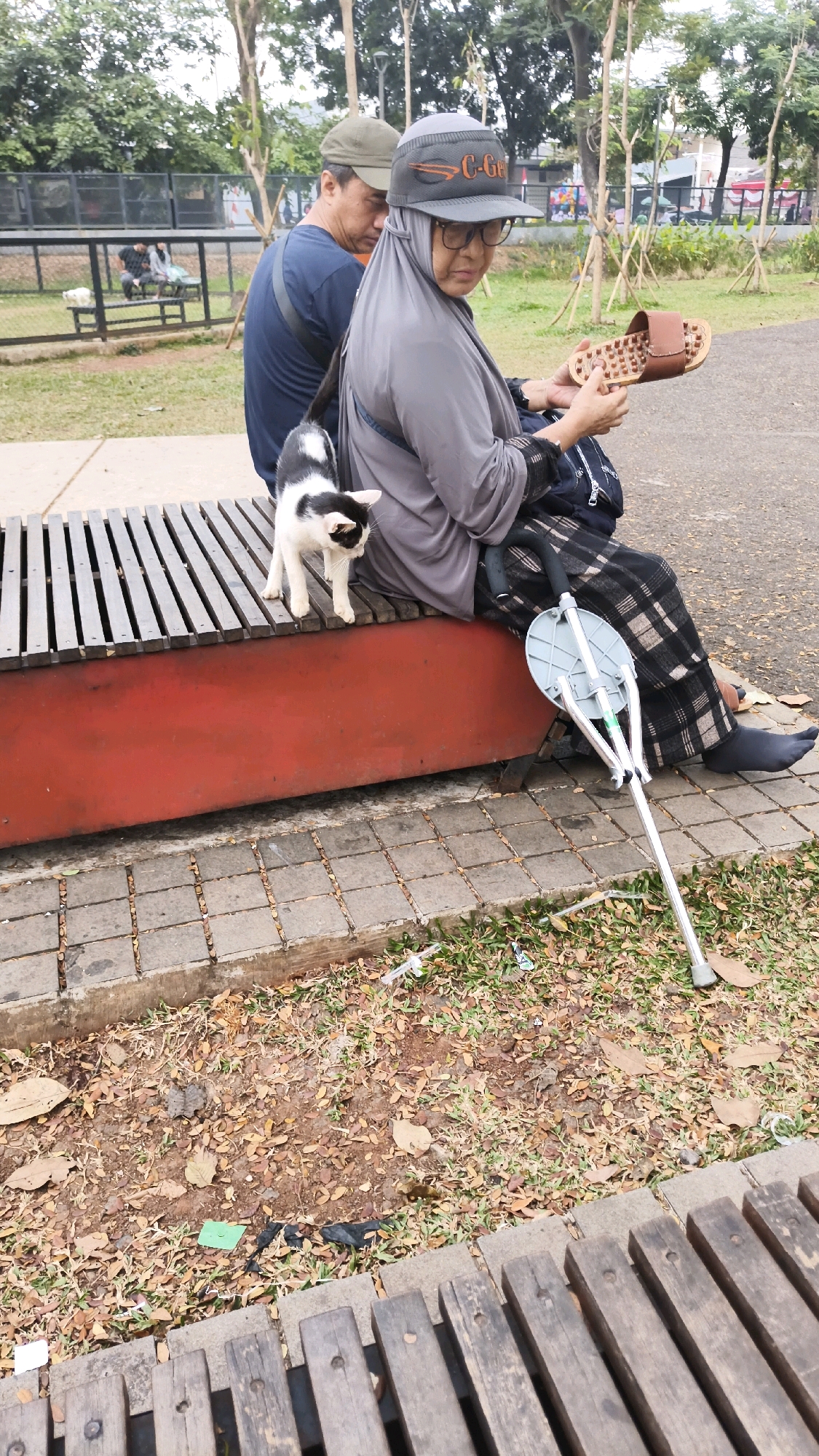 A medium shot captures an outdoor scene in South Jakarta, Indonesia, focusing on two individuals sitting on a park bench. On the left, a person wearing a green cap and a navy blue t-shirt is partially visible, looking towards the right. To their right, a woman in a grey headscarf and glasses is seated, holding a brown sandal in her right hand. She is wearing a plaid skirt and dark socks. A black and white cat stands on the wooden slats of the bench between the two people, looking towards the right. A foldable crutch with a grey seat attachment rests on the ground to the right of the woman. The bench has a red base and wooden slats. The ground in front of the bench is covered with patchy grass, fallen leaves, and some litter, including a green plastic bag. In the background, trees and fencing are visible, suggesting a public park or recreational area.