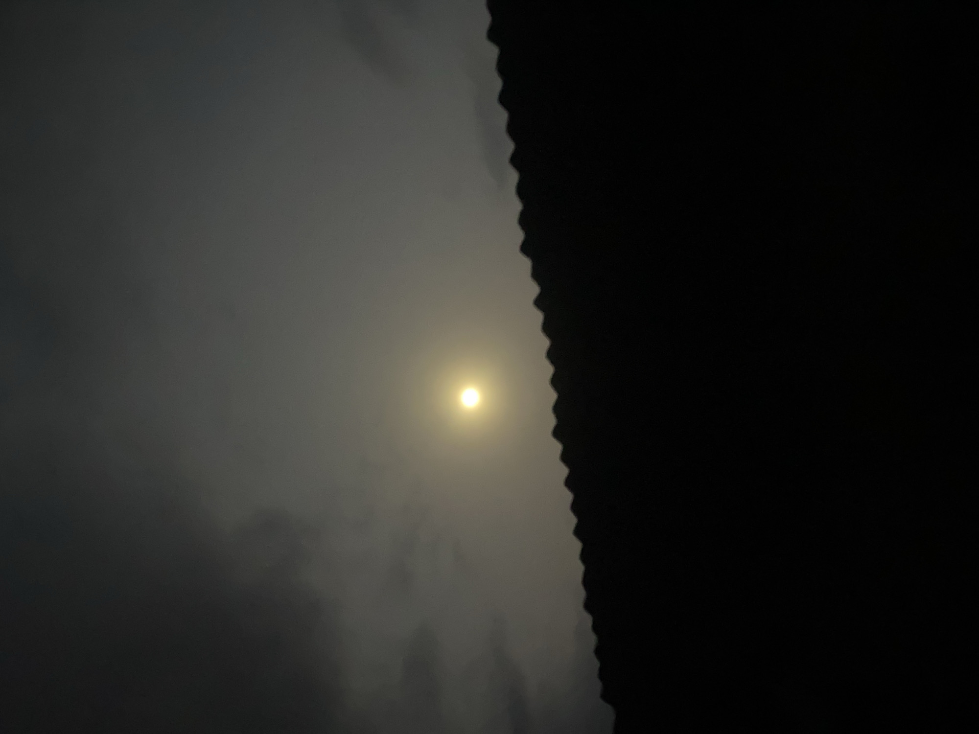 This vertical image captures a nocturnal scene, likely from Igbara, Nigeria, focusing on a bright, diffuse light source, presumably the moon, partially obscured by clouds or mist. The moon appears as a luminous, circular glow, its yellow-white radiance softened and spread by the surrounding atmospheric haze. Darker grey clouds are visible around the light source, creating a textured, obscured sky across the left and central parts of the frame.
A sharp, dark, and distinctly jagged silhouette runs vertically along the entire right edge of the image. This element, which appears to be a structural feature like a corrugated rooftop or a patterned wall, creates a strong contrast against the hazy sky. The transition from the illuminated sky to the absolute blackness of this silhouette is abrupt and distinct. The overall impression is one of natural beauty at night, with the moon peeking through a cloudy veil, framed by an architectural element. There are no people, explicit activities, or visible text in the image. The time of day is clearly night, and the weather appears hazy or misty.