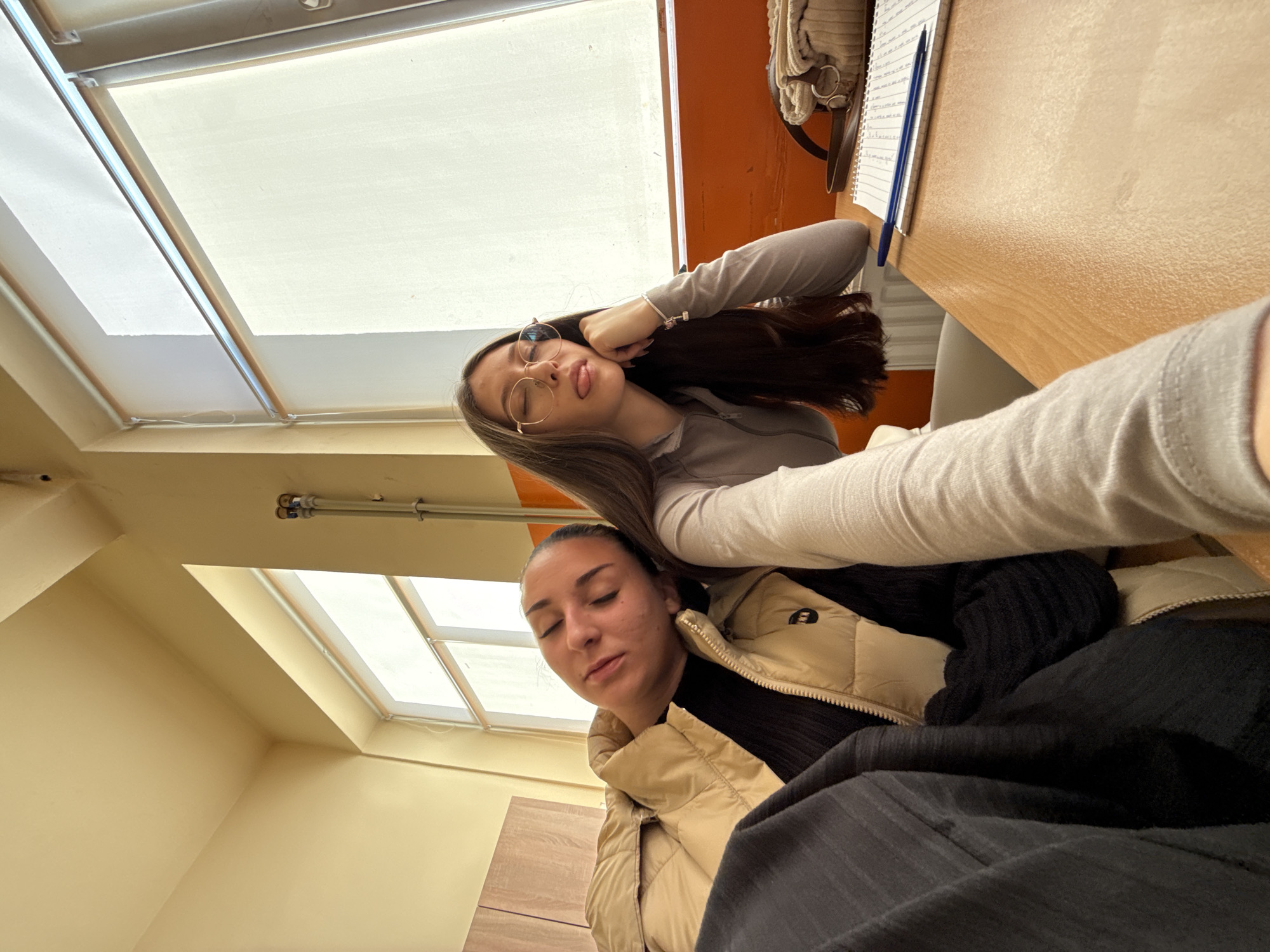 Two women are in a room. One woman is wearing glasses and has brown hair. She is holding her hair and sticking out her tongue. The second woman is wearing a beige jacket and black shirt. She has her eyes closed. A window with a white shade is in the background. A desk with a notebook and pen is visible. The room appears to be indoors, likely a classroom. The city of Sofia, Bulgaria is not determinable from the image.