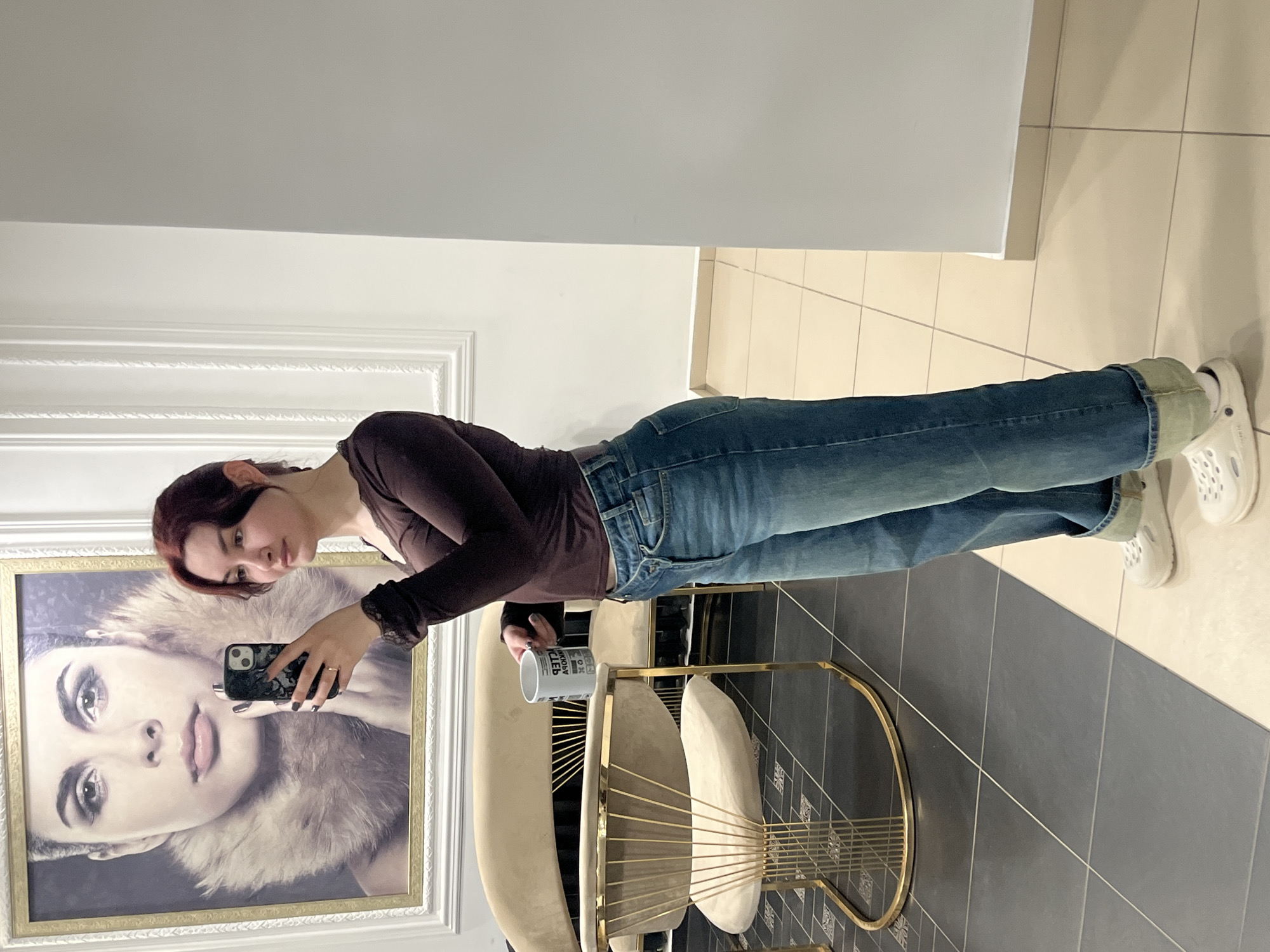 A young woman is depicted standing indoors in an establishment located in Neftekamsk, Russia. She is positioned facing slightly to her right, holding a dark-colored smartphone in her right hand, angled as if taking a selfie in a mirror or reflective surface not fully visible. Her left hand holds a white mug with black Cyrillic text, partially legible as "ДРЭМ". The woman has reddish-brown hair, light skin, and is wearing a dark brown long-sleeved top with lace cuffs, blue denim jeans featuring a distinct criss-cross front closure, and white clogs. A gold-toned ring is on her right ring finger, and her fingernails are painted dark.
The background on the left features a large, ornate gold-framed portrait on a white wall, depicting a woman with dark hair, prominent facial features, and a light-colored fur collar. Below the portrait, white decorative wall molding is present. The floor transitions from dark grey square tiles on the left to lighter beige square tiles on the right. In the mid-ground, behind the woman, a curved beige velvet sofa and a matching armchair with an intricate gold-colored metal base are visible. The walls in the immediate vicinity are white and unadorned.