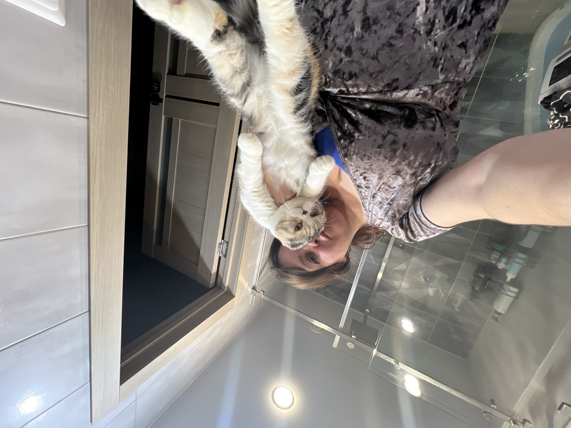 A woman is holding a cat. The location is inside a bathroom with light grey tiles and a glass shower screen, and a door with a light wooden frame. No location can be confirmed from the information in the image.