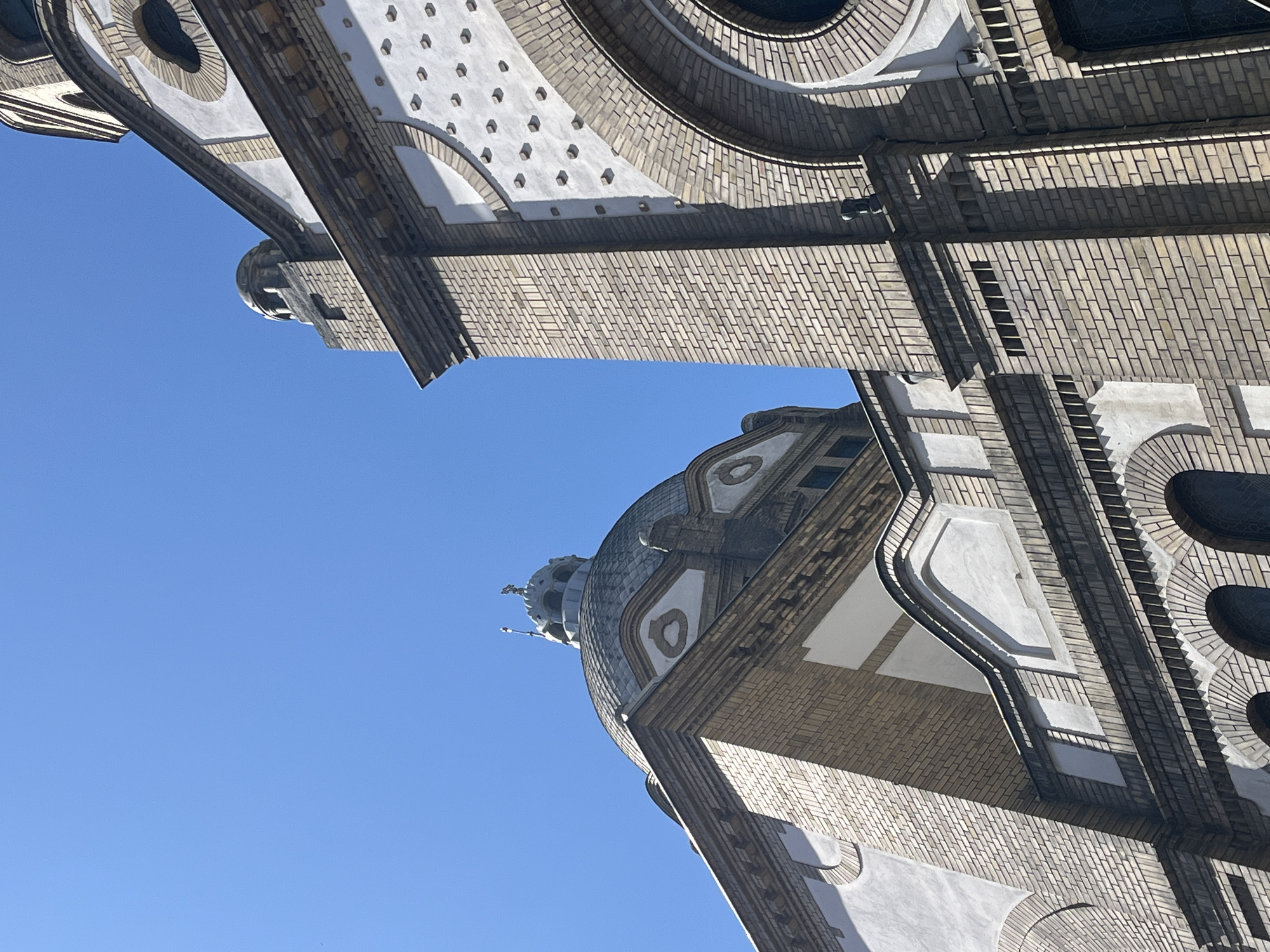 The media file displays a low-angle, upward-looking view of a grand architectural structure, likely a church or cathedral, set against a clear blue sky. The image is rotated 90 degrees counter-clockwise, showing the building primarily on the right and bottom, with the sky filling the upper and left portions.
The main subject is a large, ornate building characterized by its extensive use of light-colored brickwork (tan or beige tones) laid in detailed patterns. Prominent features include a large dome, crowned by a cupola and a finial at its apex, visible towards the lower-center. The building is adorned with decorative white or light grey stone and stucco elements, including numerous arches, circular windows or decorative motifs with radiating brick surrounds, and recessed panels. Several deep, dark circular windows are visible, suggesting either stained glass or significant depth.
Strong, bright sunlight casts sharp, well-defined shadows across the building's surfaces, emphasizing its texture and three-dimensional form. This indicates a clear, sunny day with no clouds in the bright blue sky. A section of what appears to be a bell tower or a similar tall architectural element is also visible in the upper left, constructed with matching brickwork and decorative arches.
No people or specific events are depicted; the image focuses solely on the building's architecture. The style suggests a historical European structure, possibly late 19th or early 20th century, consistent with buildings found in Central or Eastern Europe. There is no visible text in the image. The location context provided is Novi Sad, Serbia.