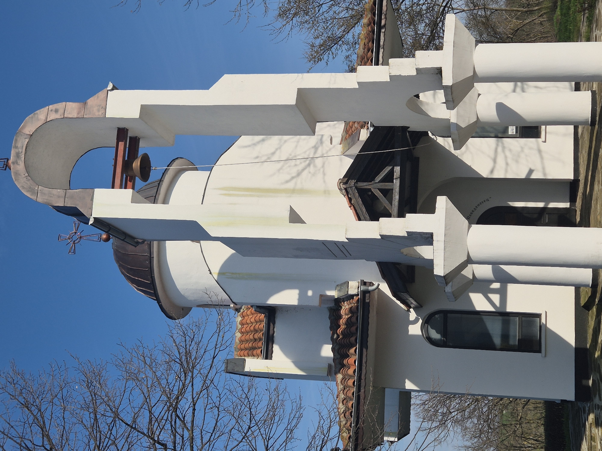 The image depicts the church "St. Nicholas the Wonderworker" in Shumentsi, Bulgaria. The church features white walls, a copper dome topped with a cross, and a bell tower with a bell. The architecture includes a series of arched openings and columns. The scene is captured on a bright day with a clear blue sky and bare trees in the background.