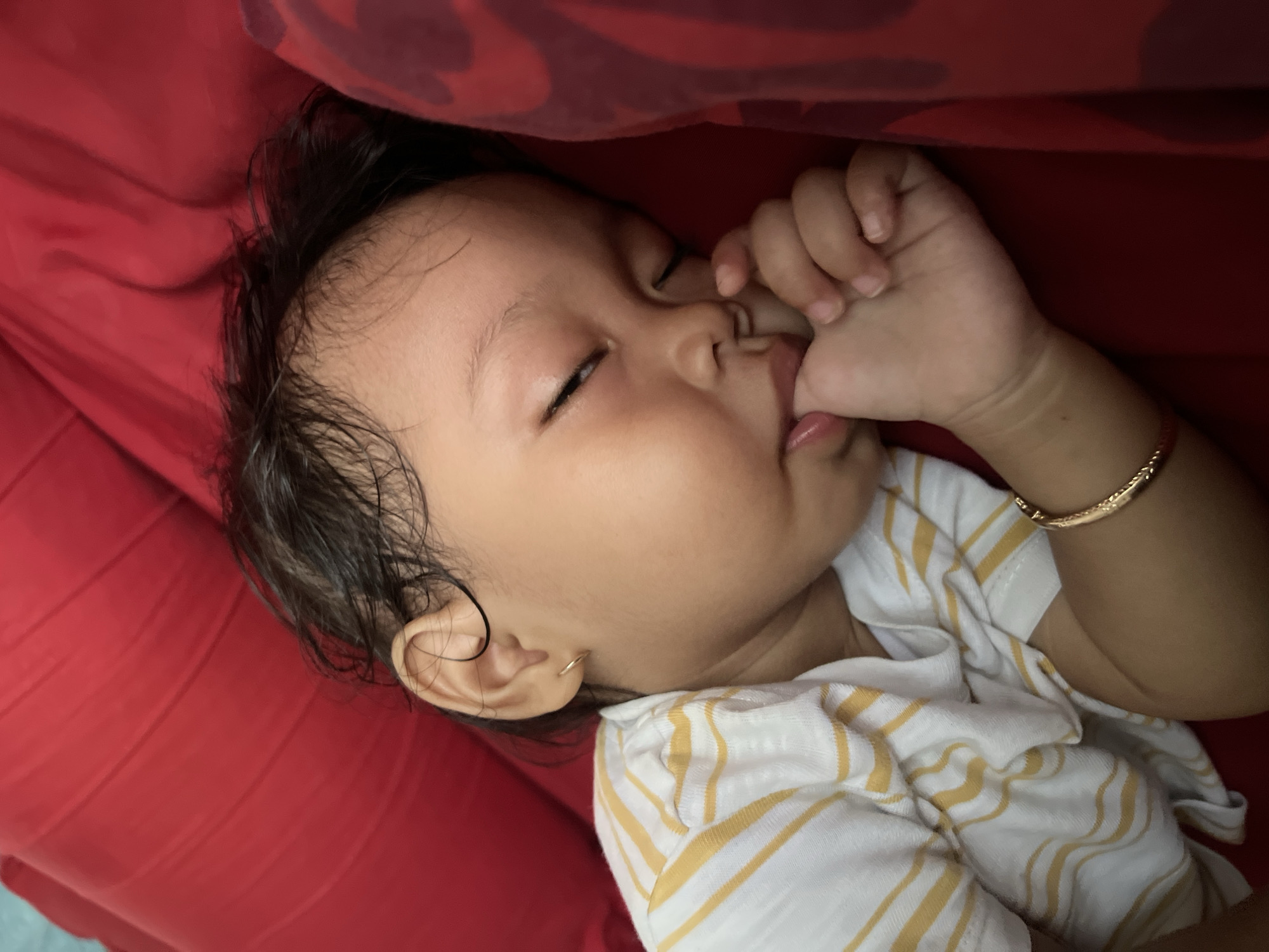 A close-up, overhead shot shows a young child sleeping on a red surface, likely a bed or couch. The child, with dark, curly hair, is lying on their side, with their eyes closed and thumb in their mouth. They are wearing a white and yellow striped shirt and a gold bracelet on their wrist. The red surface appears to be made of a soft material, possibly fabric. The lighting is soft and natural, creating a peaceful atmosphere. The image is undated and unsourced, so its exact location in Kecamatan Pati, Indonesia, cannot be determined.