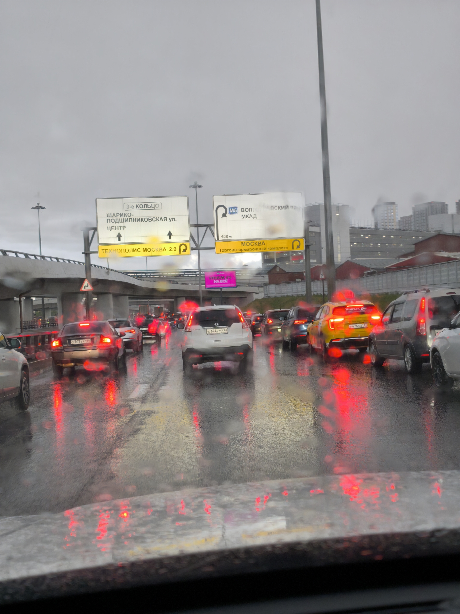The image captures a traffic jam in Moscow, Russia, on a rainy day. Multiple cars are congested on a multi-lane road under an overpass. The road surface is wet, reflecting the red tail lights of the vehicles, including a Yandex Go taxi. Overhead road signs indicate directions to the 3rd Ring Road, Shariko-Podshipnikovskaya Street, the city center, and the Volgogradsky Prospect/MKAD, identifying this road as part of the M5 highway. A sign points to "Technopolis Moscow" 2.9 kilometers away. Buildings are visible in the background, suggesting an urban environment.