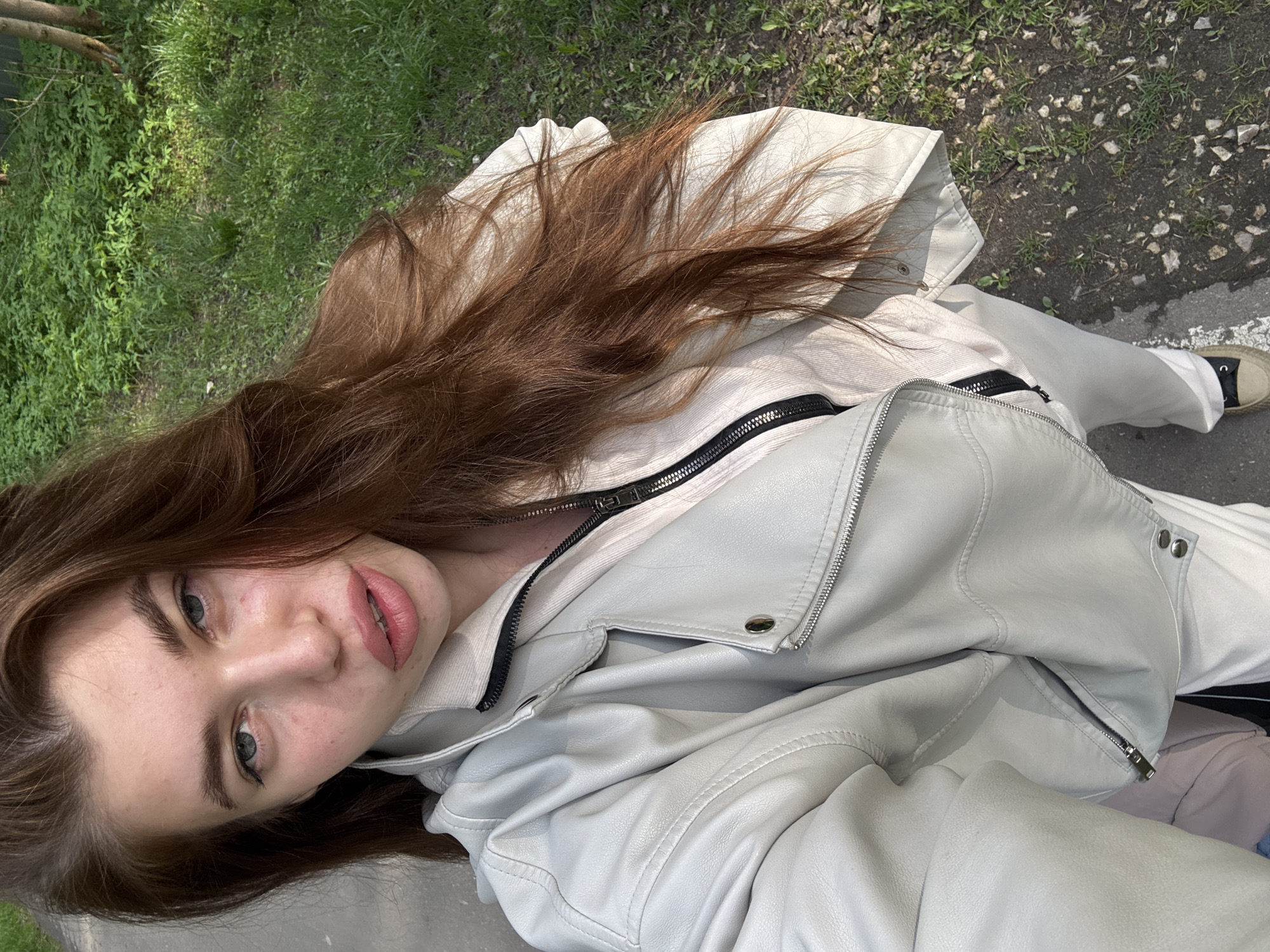 A young woman with long brown hair and green eyes is depicted from an overhead angle, likely a selfie. She wears a light-colored leather jacket with visible zippers. The background shows a mix of green grass and a paved surface, suggesting an outdoor urban environment. Her face is visible, with emphasis on her eyes and lips. The image is taken in natural light, giving it a casual, candid feel. There are no apparent signs indicating the location is in Мещёры, Russia.