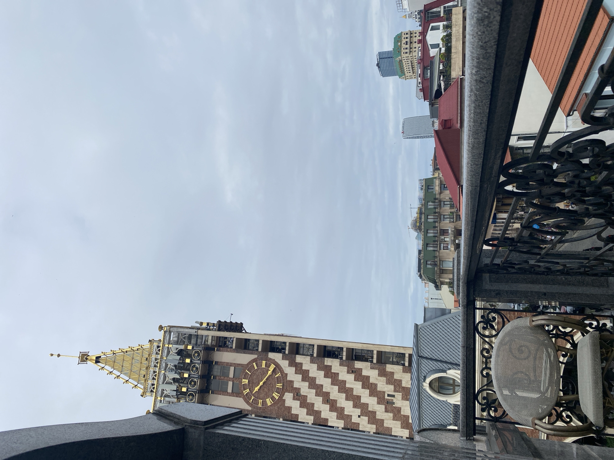 This is a medium-angle shot of the Batumi Clock Tower, taken from a balcony overlooking a city skyline under an overcast sky. The tower, predominantly red and cream colored with a distinctive zig-zag pattern, features a large, golden clock face with Roman numerals. The upper section of the tower is gilded, with a spire at its apex. In the foreground, the granite railing of a balcony is visible, along with an ornate black metal balustrade. A wicker chair with a cushion sits on the balcony. Behind the clock tower, various other buildings are visible, including some with red roofs and a modern, tall blue skyscraper. The sky is filled with thick, gray clouds.