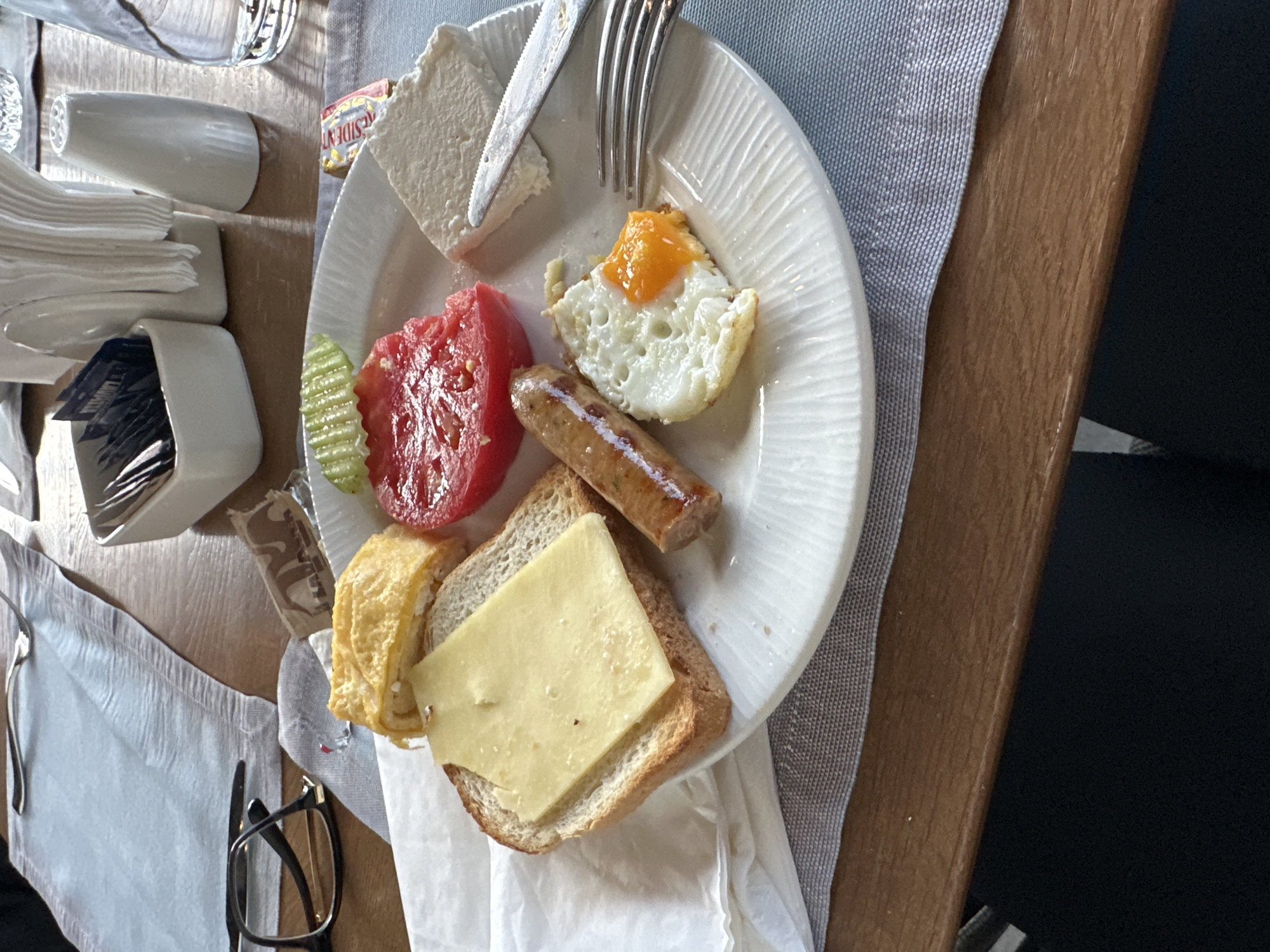 A top-down shot shows a plate of food on a table, likely a breakfast. The plate contains a piece of feta cheese with a knife, a fried egg, a slice of tomato, a sausage, a slice of bread topped with a slice of cheese, and a rolled omelet. Other items on the table include a napkin holder with napkins and packets of sugar or sweetener, a glass, eyeglasses, silverware, a butter packet, and a placemat. The table is made of wood, and a part of a chair is visible on the right. The environment suggests an indoor setting, possibly a restaurant or dining area. The city and location within Kazanlak, Bulgaria, are not deducible from the image.