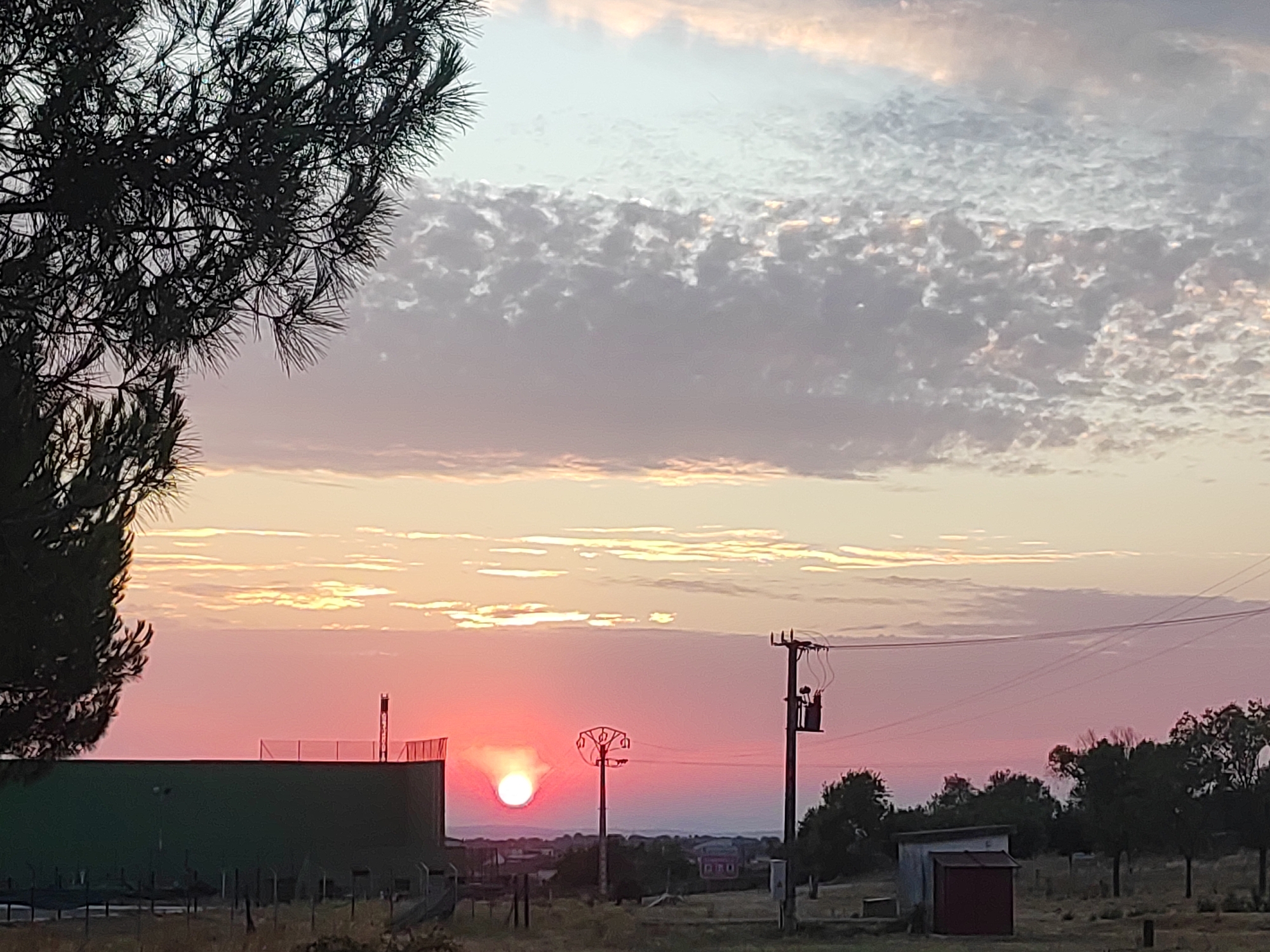 This media file depicts a sunset scene in Sobradillo, Spain. The sky is filled with a gradient of colors, transitioning from a soft pink and orange near the horizon to a pale blue at the top, with scattered, textured clouds. A bright, circular sun is low on the horizon, casting a warm glow and radiating light.
In the foreground, a large, dark silhouette of a pine tree branches into the upper left corner. On the right side of the image, a wooden utility pole with electrical equipment stands prominently, with wires stretching across the sky. To the left of the pole, a tall, slender structure with a circular top is visible. Further left, a long, dark green building with a fenced top is present. In the distance, a rural landscape with scattered trees and buildings can be observed. A small, rustic shed with a red door is situated in the lower right portion of the image, with dry grasses and sparse vegetation covering the ground.
The overall mood is tranquil and serene, characteristic of a peaceful sunset. The time of day is clearly evening, as indicated by the setting sun. The weather appears clear, with the sun breaking through the clouds. No text is visible in the image.