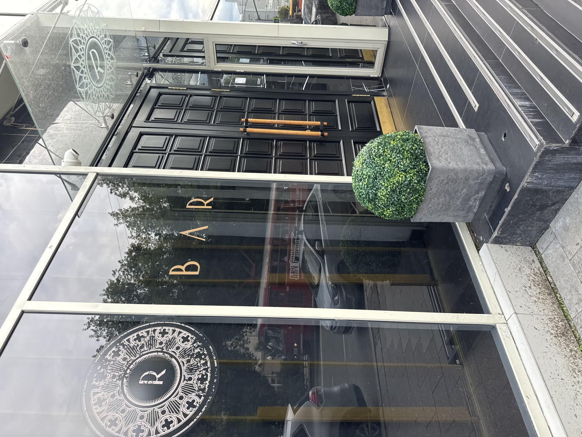 The image shows the exterior of a building, likely a bar or restaurant, in Yekaterinburg, Russia. The entrance features double black doors with wooden handles and a large glass facade. The word "BAR" is visible in gold lettering on the glass. A circular emblem with the letter "R" in the center is also present on the glass, repeated on a decorative panel above the doors.
A decorative spherical bush in a concrete planter sits to the right of the entrance. To the right of that, a set of stairs with metallic trim ascends. The building's facade is modern, with black tiles and reflective glass. The reflection in the glass shows trees and a cloudy sky, suggesting it is daytime and potentially overcast weather.
The image appears to be taken from a slightly elevated, ground-level perspective, looking towards the entrance of the establishment. There are no people visible in the immediate foreground or on the entrance. The scene is neat and well-maintained, giving a sense of a commercial establishment. The presence of the "R" emblem and the word "BAR" are the most prominent text elements.