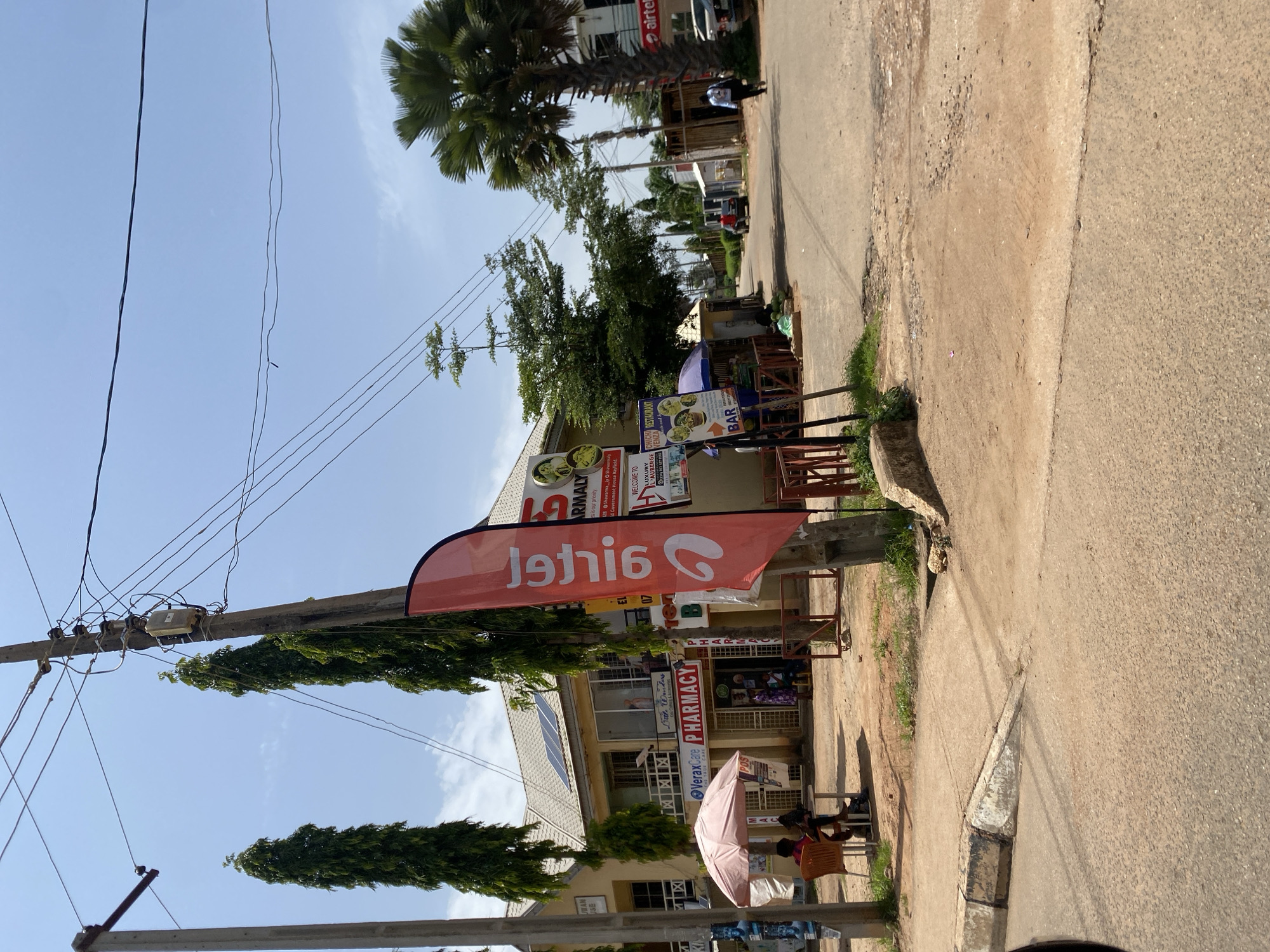 The image depicts a street scene in Bar Jirgi Summa, Nigeria. A building with a light yellow facade and a tiled roof, possibly containing a pharmacy and other businesses, is visible. Solar panels are installed on the roof. In front of the building, there are several storefronts, including "VeraxCare Pharmacy" and a store with a pink umbrella and seating outside. A red banner with white text in a reversed font is prominently displayed on a pole.
The street is paved and appears to be a main thoroughfare, with other buildings and structures lining it. A large palm tree stands on the right side of the image, near an Airtel advertisement. Several utility poles with numerous wires crisscross the sky. A few people are visible in the scene, including individuals sitting at tables outside one of the establishments and others walking or driving on the street. The environment suggests a warm, sunny day, with clear blue skies. The overall impression is one of a bustling commercial area in an urban or semi-urban setting.