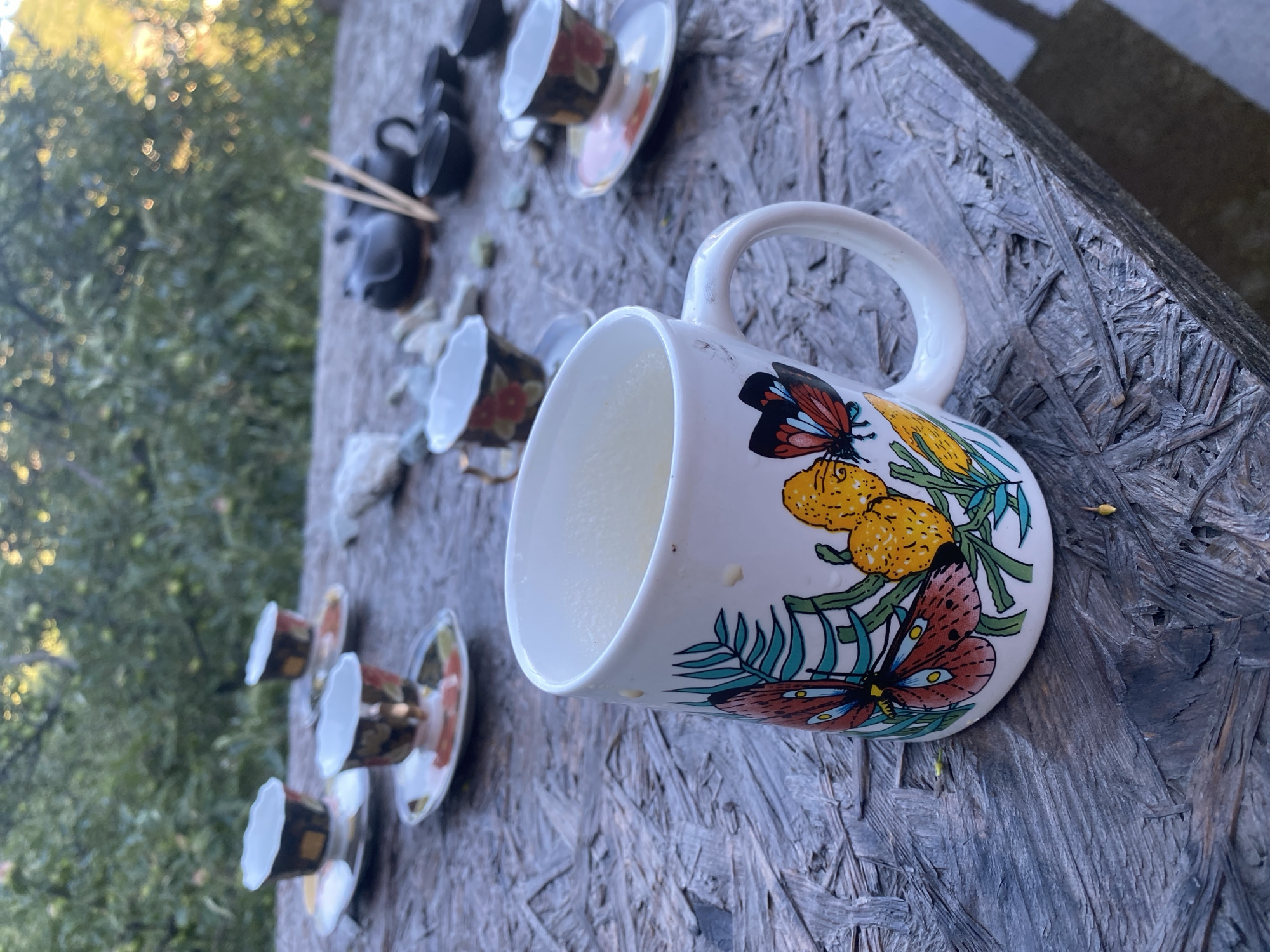 The image shows a close-up view of a white mug decorated with colorful butterflies and flowers, placed on a rustic wooden table outdoors. The mug is filled with a light-colored liquid, likely a beverage. Several small teacups and saucers with floral designs are scattered around the table, along with a dark-colored teapot and chopsticks, suggesting a tea or coffee setting. In the background, lush green foliage and trees are visible, indicating an outdoor, possibly garden or park, setting. The sunlight filtering through the trees suggests it is daytime. There is no indication of people or any specific location in Almaty, Kazakhstan.