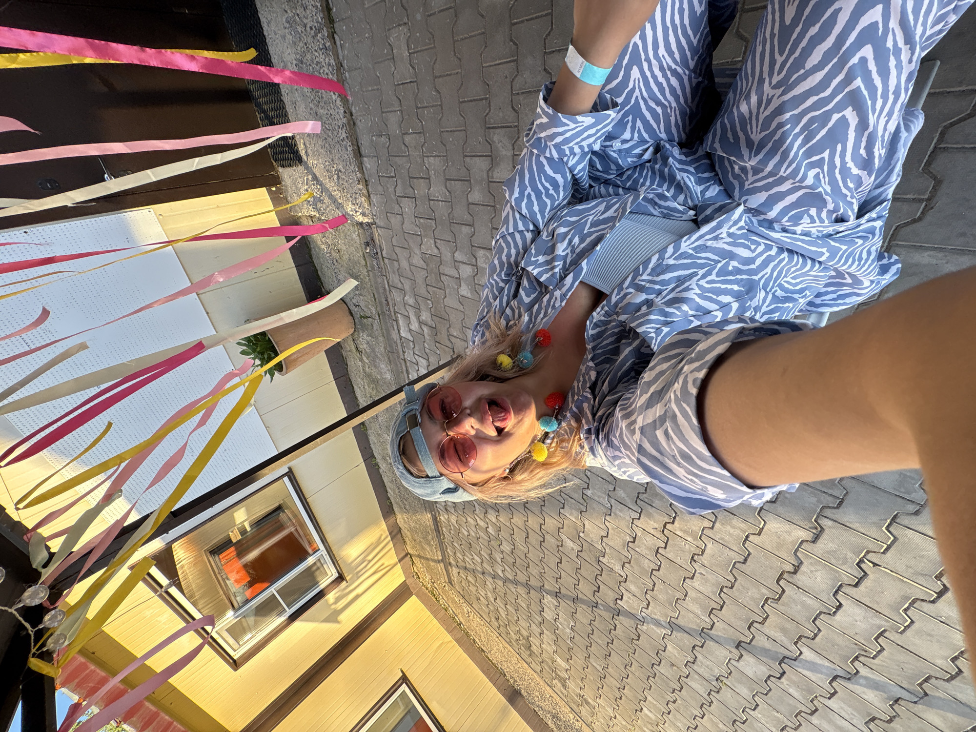 A young woman with blonde hair and pink sunglasses is sitting on a paved area, with her tongue sticking out. She is wearing a blue and white zebra-print outfit, a blue baseball cap, and colorful pom-pom earrings. A turquoise wristband is visible on her right arm. Above her, colorful ribbons are hanging, suggesting a festive atmosphere. The background includes a yellow building with a window and a dark door. The scene appears to be outdoors, possibly in a yard or patio area, under natural sunlight. There is no discernible information in the image to connect it to Birobidzhan, Russia.