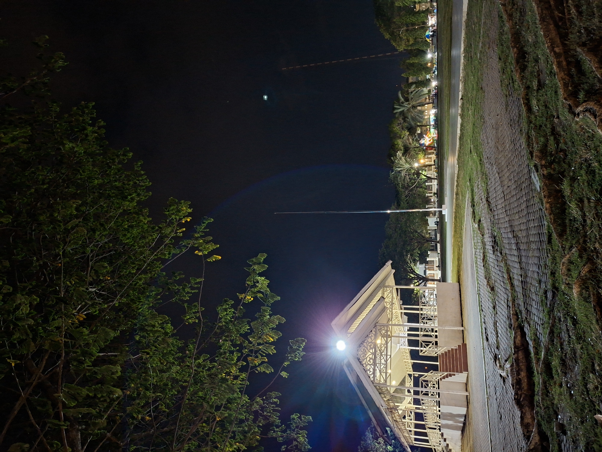 This media file captures a vibrant night scene in Lhokseumawe, Indonesia, shown in a vertical orientation but depicting a landscape. The time of day is clearly night, with a dark sky illuminated by various artificial light sources.
In the mid-ground, a prominent, elegant white pavilion or gazebo stands, featuring intricate lattice work and railings. A bright lamp mounted on this structure emits a strong light, creating a starburst effect and lens flare, illuminating parts of the structure and the surrounding foliage. Steps lead up to the pavilion.
To the foreground and left, lush green trees and bushes with dense foliage are partially lit by the pavilion's light, creating areas of deep shadow and bright highlights. Some of the leaves appear yellowish, possibly due to the artificial lighting or seasonal changes.
Stretching horizontally across the image, beyond the pavilion, is a paved pathway or road. To the right of this path, there's an expanse of patterned paving, possibly cobblestones or bricks, interspersed with patches of grass and earth. Further in the background, a row of diverse trees, including tall palm trees, lines the distant edge of the path. Behind these trees, multiple illuminated buildings and what appear to be stalls or structures with various colorful lights are visible, suggesting a bustling area, possibly a night market or recreational space.
The upper portion of the image is dominated by the deep black night sky. A faint, thin light streak, possibly an aircraft contrail or light trail, can be seen high in the sky, along with another small, bright light spot. No specific human activity is explicitly captured, but the illuminated setting implies a lively public area for evening leisure or events. There is no visible text in the image. The overall mood is one of a serene yet active tropical night.