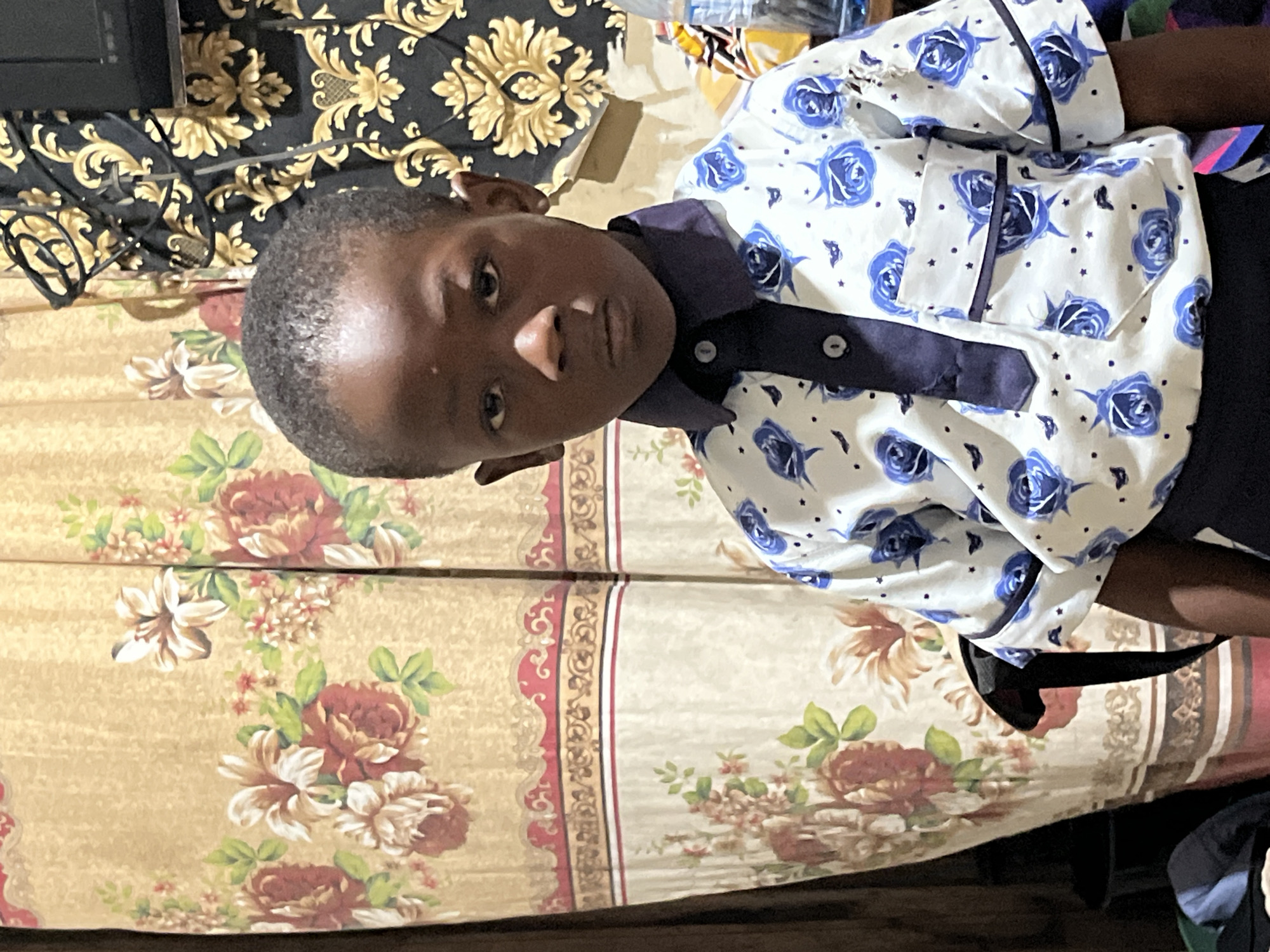 A young boy poses for a portrait on a bed or mattress with floral bedding. He is wearing a white shirt with blue rose patterns and a dark blue collar and buttons. Part of a black and gold patterned surface (possibly a headboard or wall) is visible in the upper left corner of the frame. The context of the image suggests the location is Badagry, Nigeria.