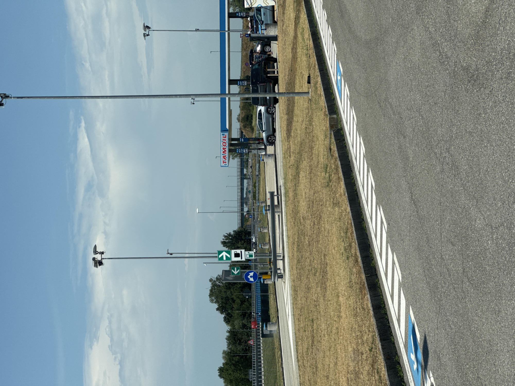 This is a photograph of a service station on a sunny day. In the background, a TAMOIL gas station is visible with several cars parked and refueling. The service station appears to be located in the city of Occimiano, Italy, indicated by the presence of Italian road signs. The foreground shows a portion of an asphalt road with white stripes and a blue handicapped parking symbol. The environment is characterized by dry, brown grass and scattered trees. The sky is partly cloudy.