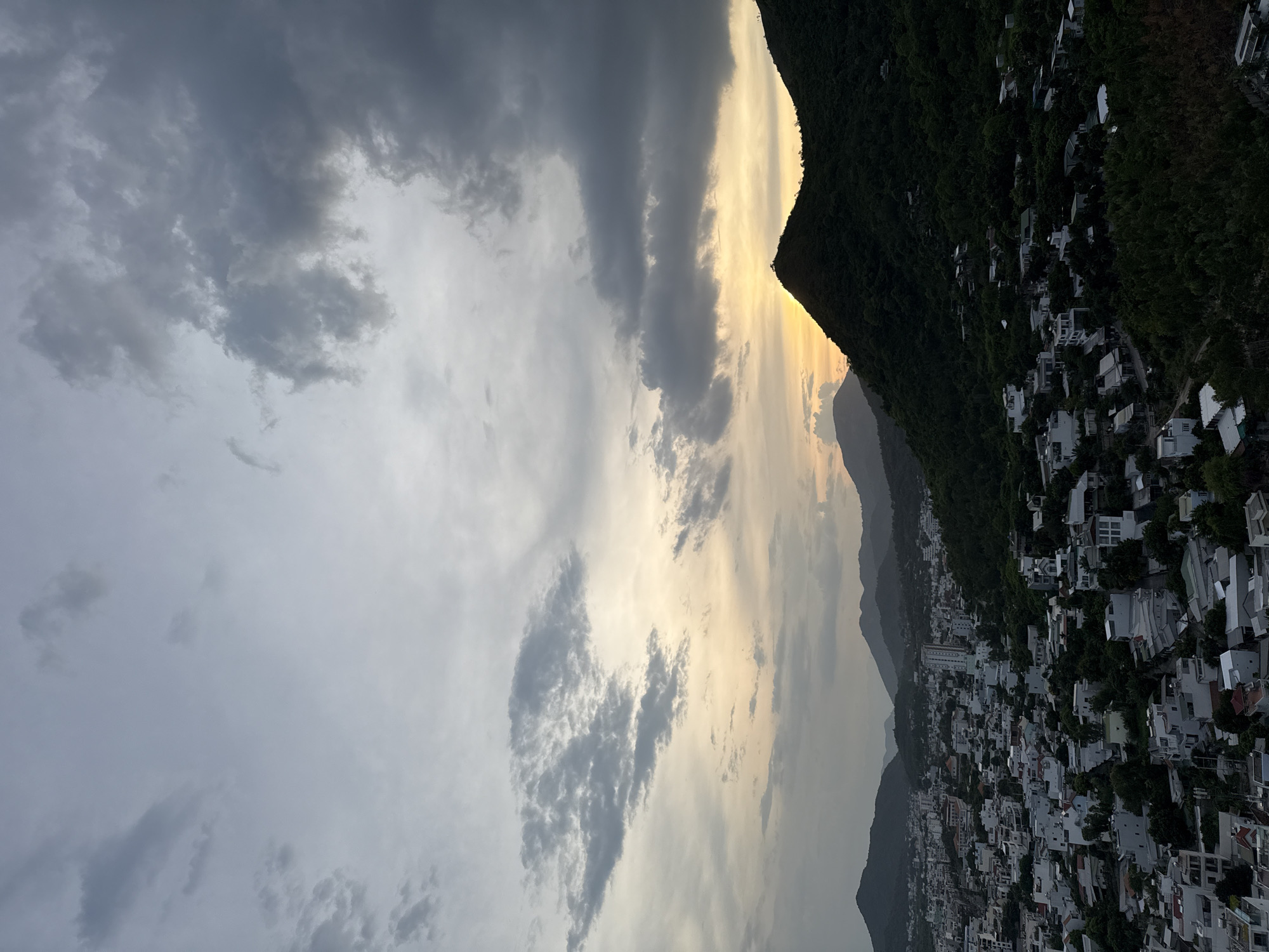 This image captures a sprawling hillside town nestled beside a densely forested mountain range, under a dramatic sky. The town, characterized by its clusters of white-roofed buildings, cascades down the steep slope. The architecture suggests a modern, perhaps resort-like, development. Lush green vegetation covers the mountainside, contrasting with the urban sprawl.
The sky is a prominent feature, filled with a mix of dark, brooding clouds and patches of lighter, illuminated sky, hinting at the end of the day or an approaching storm. A sliver of warm, golden light pierces through the clouds near the mountain peak, creating a striking contrast with the darker clouds and shadowed landscape.
The time of day appears to be late afternoon or early evening, given the quality of the light and the cloud cover. The weather seems moody and overcast, possibly with a chance of rain. The location context provided places this scene within the Bac Natural Heritage Area in Trang, Vietnam, suggesting a beautiful, potentially remote, natural landscape interspersed with human development.
There are no visible people or specific objects of focus, apart from the town and the natural environment. The overall impression is one of dramatic natural beauty combined with human habitation, with the weather playing a significant role in setting the mood of the scene. The image evokes a sense of awe at the scale of the landscape and the tenacity of human settlement within it. No visible text is present in the image.