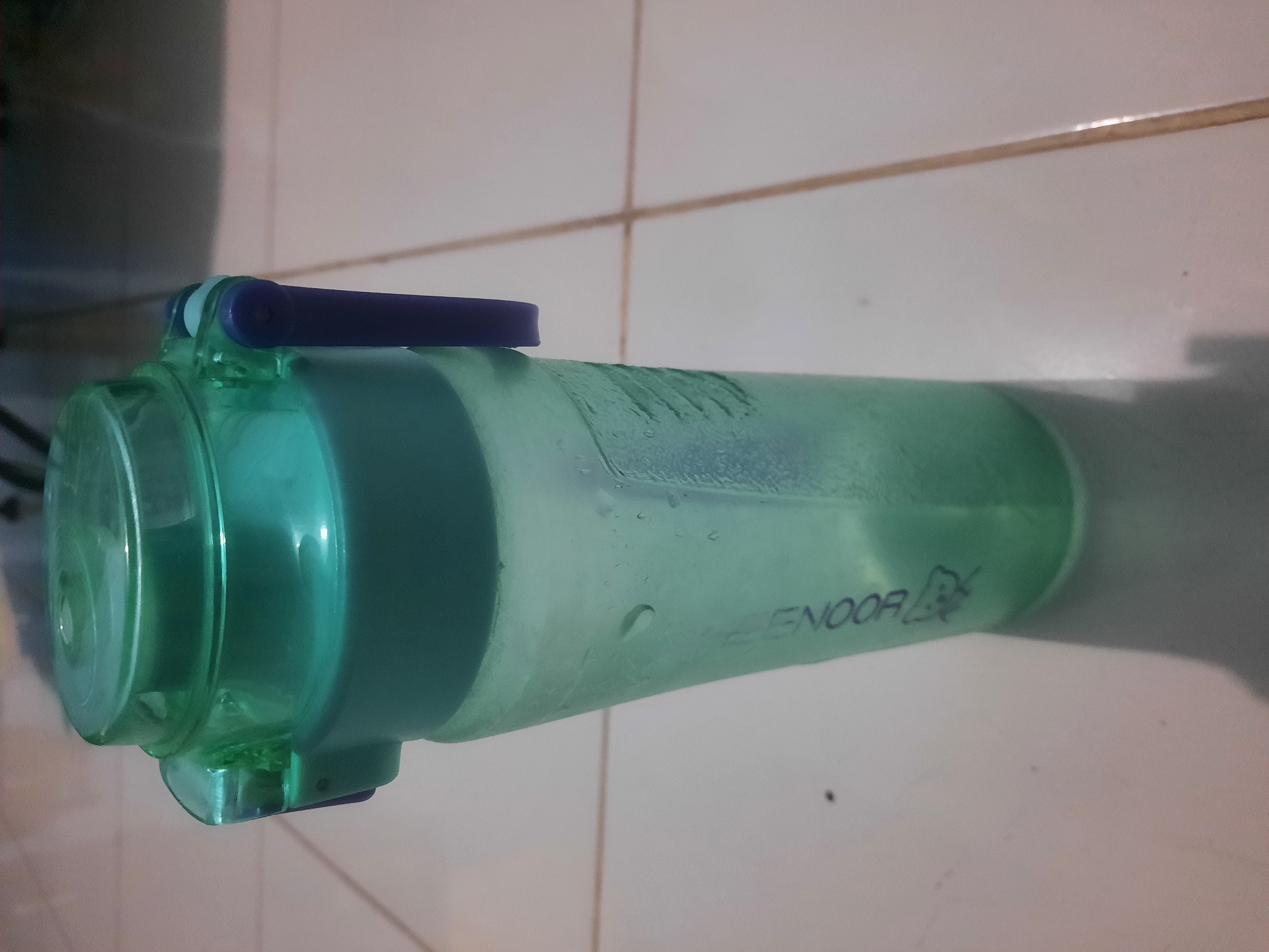 A green plastic water bottle with a blue handle is standing upright on a tiled floor. The bottle has some condensation on its surface, and text is visible on its side. The background appears to be indoors with a blurred view of what might be a wall or furniture. The scene is set in a residential area of West Jakarta, Indonesia.