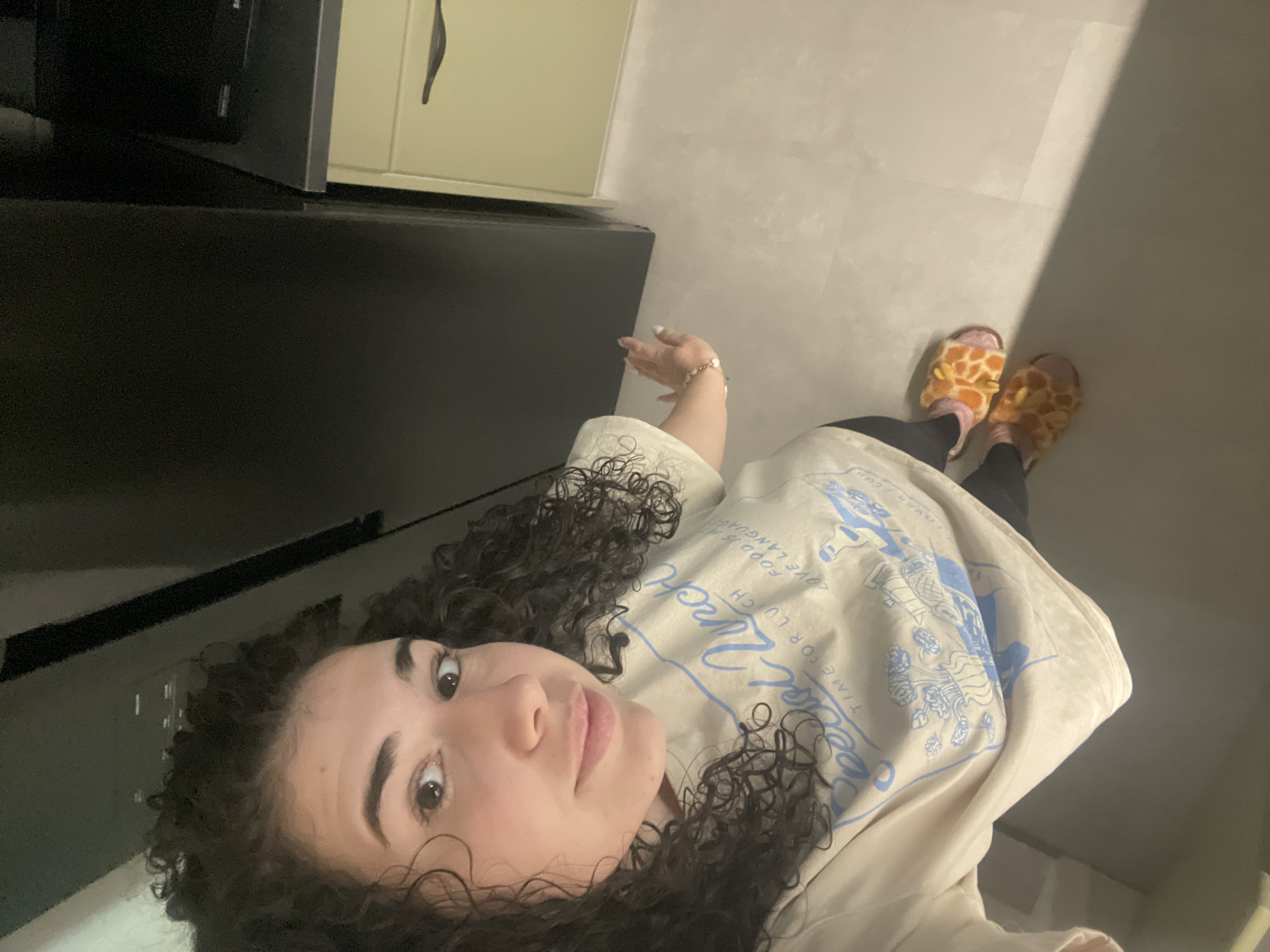 A person with curly dark hair is positioned in the lower left of the frame, looking up towards the camera. They are wearing a light-colored shirt with blue graphics. The person is also wearing black leggings and fuzzy, giraffe-patterned slippers. Their left hand is visible near the top of the frame, adorned with a bracelet.
The background consists of a black kitchen counter along the left side, a light-colored cabinet above the counter, and a light-colored floor. A bright streak of light is cast on the floor.
Given the neutral background and lack of distinct landmarks, determining the exact location within the city of Sofia, Bulgaria is not possible from this image.