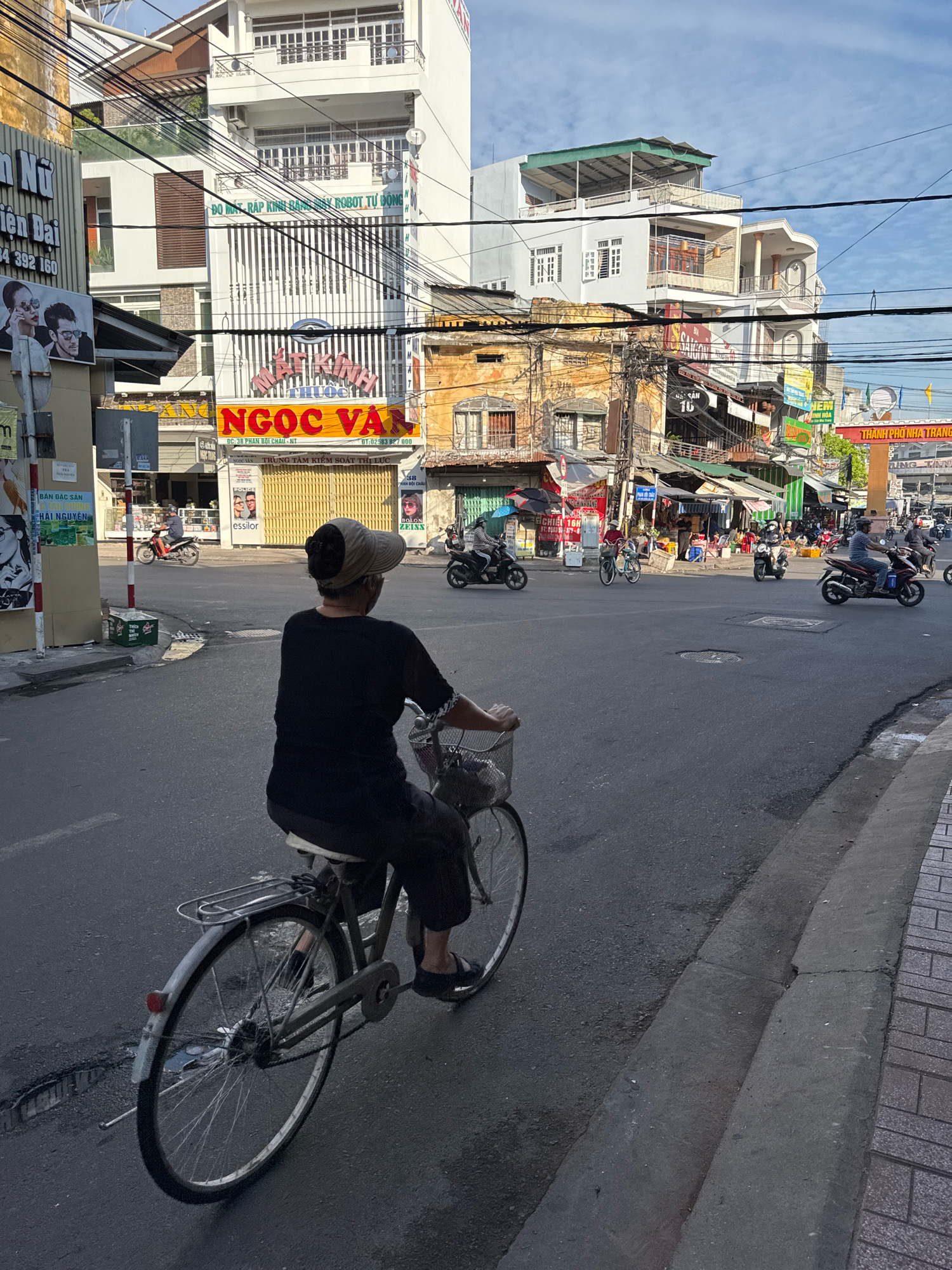 The image depicts a street scene in Nha Trang, Vietnam, characterized by its bustling urban environment. In the foreground, a person is riding a bicycle from right to left, viewed from the rear. The cyclist is wearing a dark t-shirt and dark pants, with a light-colored visor on their head. The bicycle is a standard model with a basket attached to the handlebars.
The background is filled with multi-story buildings, typical of Southeast Asian city architecture, displaying a mix of older, weathered structures and more modern ones. Many buildings feature signs with Vietnamese text, indicating businesses such as optical shops, restaurants, and other commercial establishments. The street is paved with asphalt and shows some signs of wear and tear, including cracks and potholes.
Several motorbikes are also visible on the street, some parked and others in motion, contributing to the sense of a busy urban thoroughfare. Power lines are strung overhead, a common feature in Vietnamese cities, creating a dense network against the sky. The sky itself is partly cloudy, suggesting it could be morning or late afternoon, with the sun casting shadows. The overall impression is of an active street in a Vietnamese city, likely a commercial district, with everyday life unfolding.