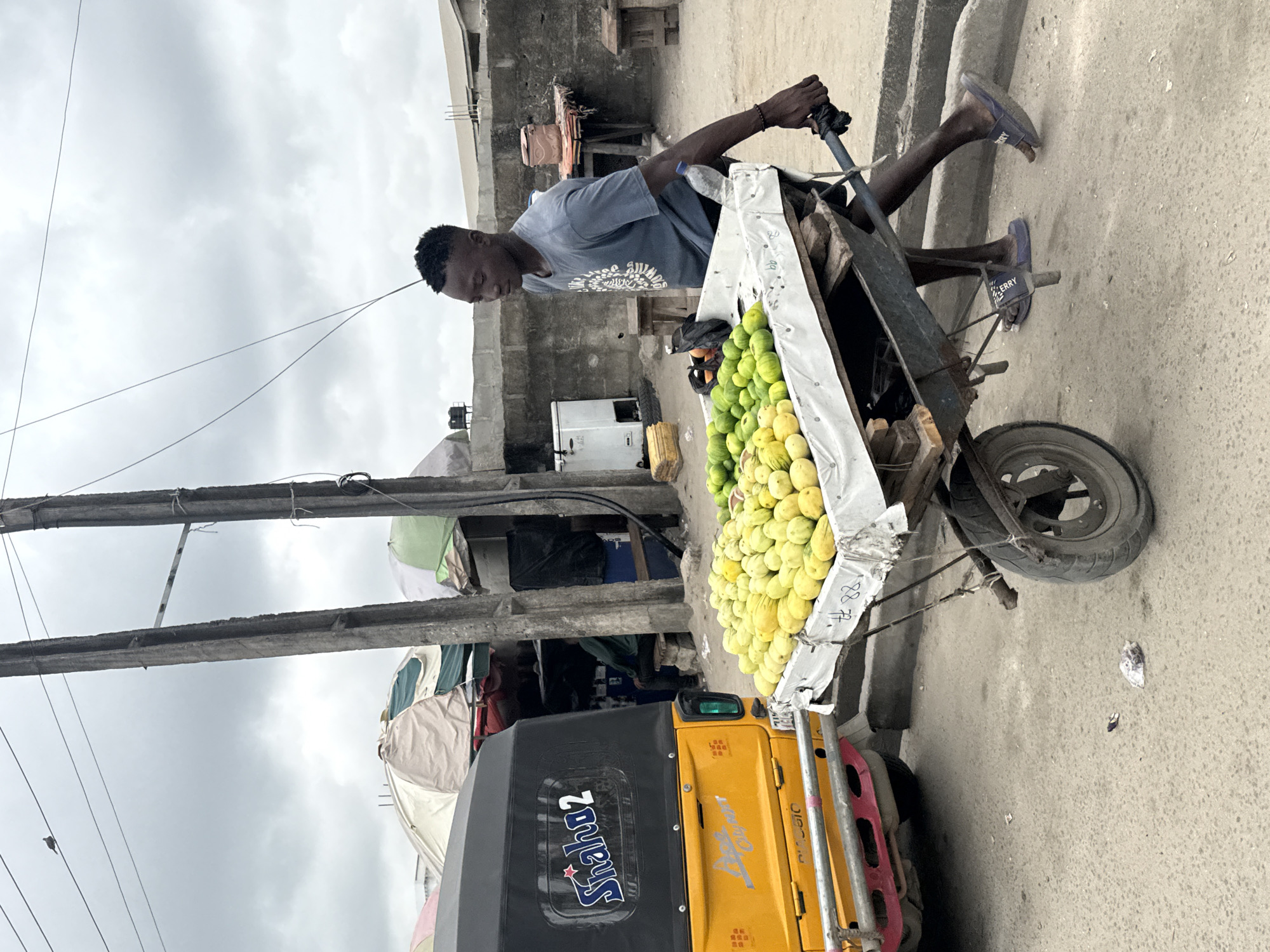 A young Black male, wearing a gray t-shirt and dark blue flip-flops, pushes a cart laden with green and yellow fruits through a dusty street in Ado, Nigeria. The cart is made of metal and wood, with a white cloth covering the top. In the background, a yellow auto-rickshaw is parked to the left. Beyond that, there are market stalls with colorful umbrellas, concrete buildings, and electrical poles. The sky is overcast, suggesting a cloudy day.