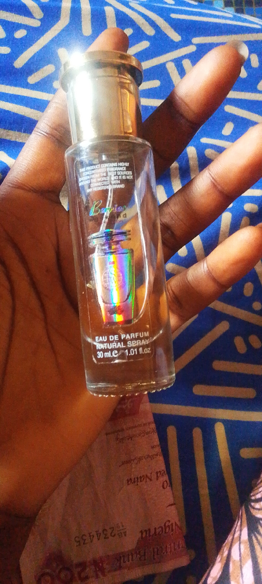 A person's hand holds a small, clear glass bottle of perfume. The bottle has a gold cap and a label with iridescent text that reads "Berries." Inside the bottle, a miniature perfume bottle with a rainbow effect is visible. Below this, text on the bottle states "EAU DE PARFUM NATURAL SPRAY 30 ml.e 1.01 fl.oz." Further text on the bottle reads, "THIS PRODUCT CONTAINS HIGHLY CONCENTRATED FRAGRANCE Drogen FROM THE BEST SOURCES AROUND THE WORLD AND IT IS NOT CONNECTED WITH ANY REGISTERED BRAND." In the background, a blue and white patterned fabric is visible. Partially obscured beneath the bottle and hand is a Nigerian 200 Naira banknote, with visible text including "Central Bank," "Nigeria," and "200." The overall scene appears to be a close-up shot with artificial lighting. The hand holding the bottle suggests a personal interaction with the product. The banknote implies the location could be Nigeria.