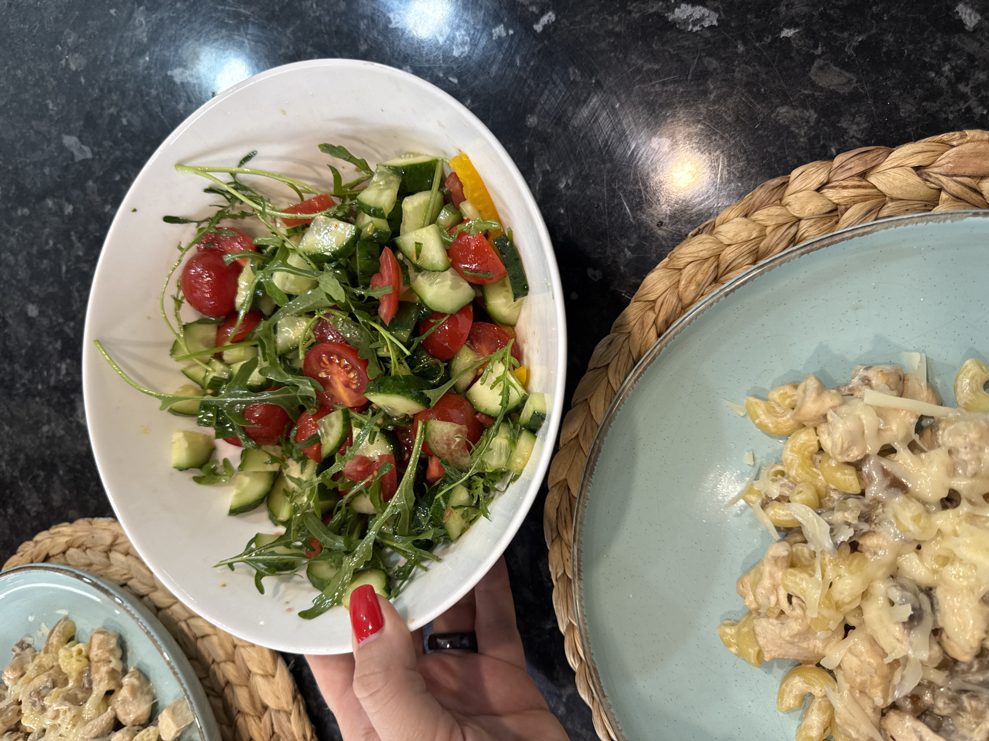 A hand with red nail polish is holding a white bowl filled with a salad. The salad consists of chopped cucumber, cherry tomatoes, and arugula. There is also a plate of pasta with chicken and cheese in the lower left corner of the image. The setting appears to be a kitchen counter. This image was not taken in Sochi, Russia.