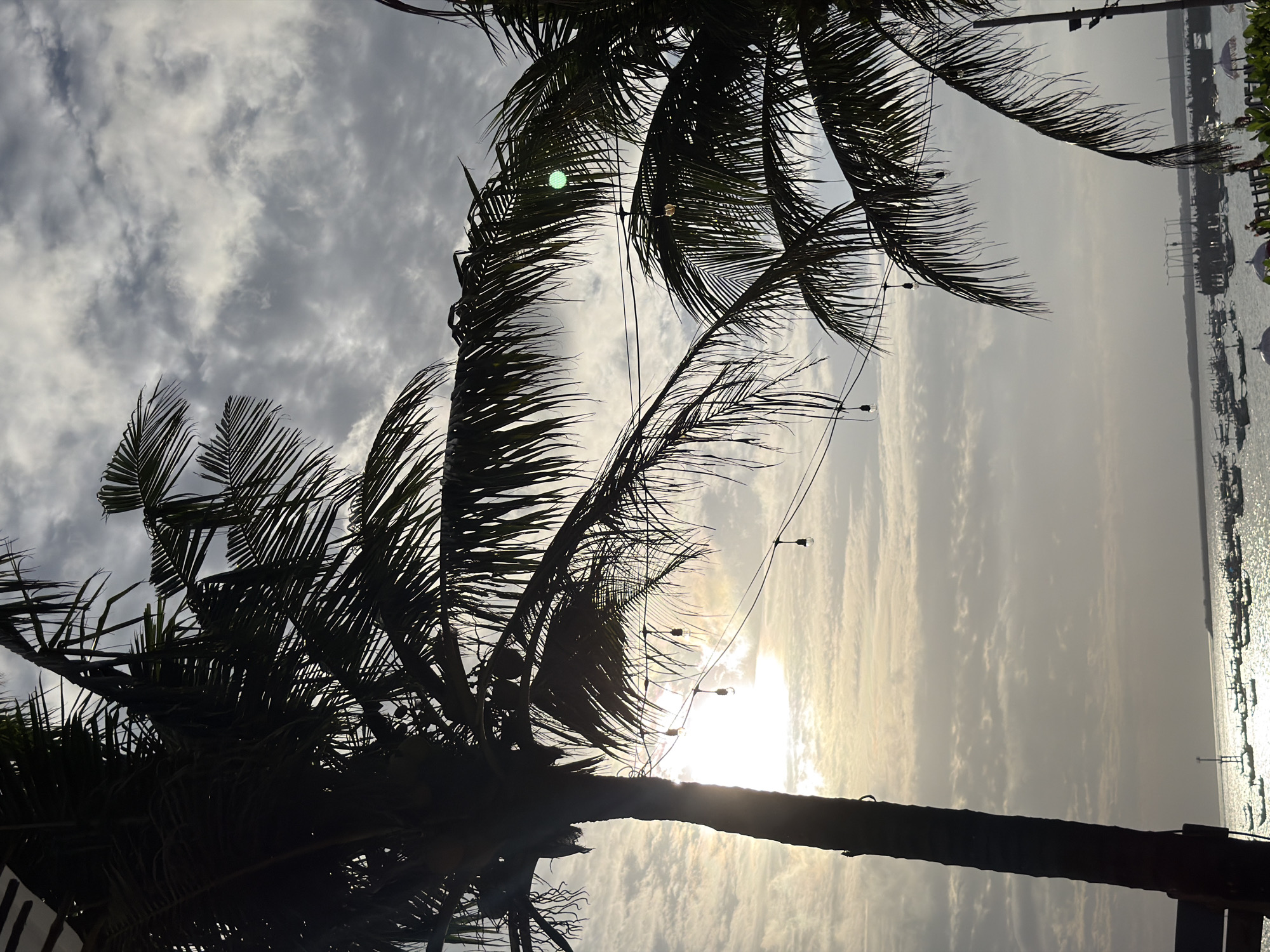 The image shows a view of the ocean in Kuta, Indonesia. In the foreground, silhouetted palm trees with their fronds are visible against a cloudy sky. String lights with exposed bulbs are strung between poles, with several lights visible. The sun is setting or rising behind the clouds, creating a soft, diffused light. In the distance, the ocean is calm, and numerous small boats are anchored near the shore. A pier or jetty extends into the water with structures and what appear to be decorative umbrellas. The overall scene suggests a coastal resort or beach area.