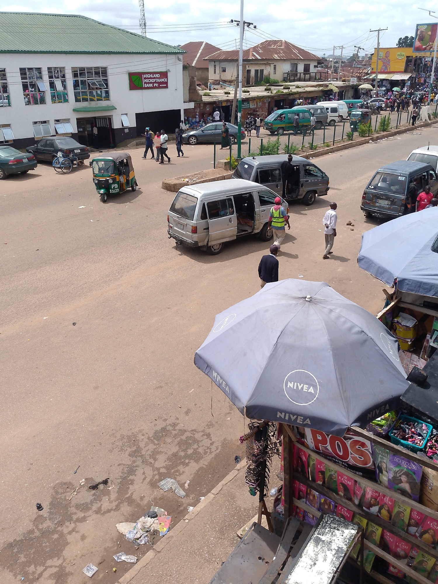 The image shows a street scene in Bukuru, Nigeria, likely a commercial area. In the foreground, two large gray umbrellas, one with "NIVEA" branding, provide shade over a small roadside stall selling various goods, including what appear to be cosmetics and hair products. Several people are visible on the street. To the left of the stall, a silver minivan with its side door open is parked. Further down the street, a green and yellow motorized rickshaw is in motion, and a silver minivan is also visible. Across the street, a white building with a green roof houses "HIGHLAND MICROFINANCE BANK PLC". Several cars and other vehicles are parked or moving on the asphalt road. The environment appears to be a typical busy urban setting with shops and market stalls lining the street.