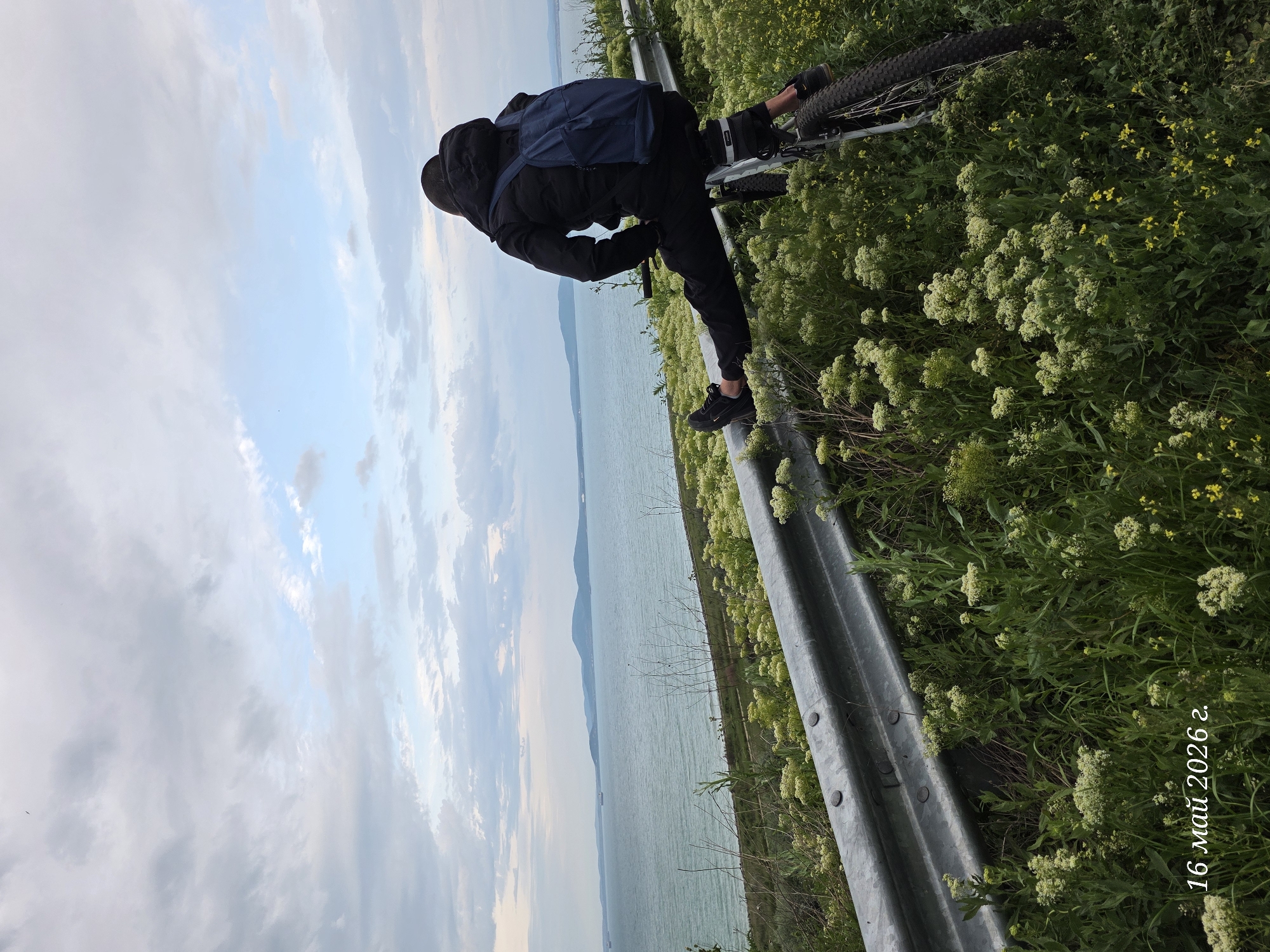 A person stands with their bicycle on a roadside overlooking a body of water. The person is dressed in black with a dark blue backpack. The bicycle is visible on the right side of the image, partly obscured by greenery. A metal guardrail runs diagonally across the scene, separating the person and bicycle from the water view. The body of water stretches into the distance, with land visible on the opposite shore. The sky is filled with clouds. The date "16 май 2026 г." is visible in the lower right corner. Based on the landscape, the scene could be located in a coastal area near Burgas, Bulgaria.