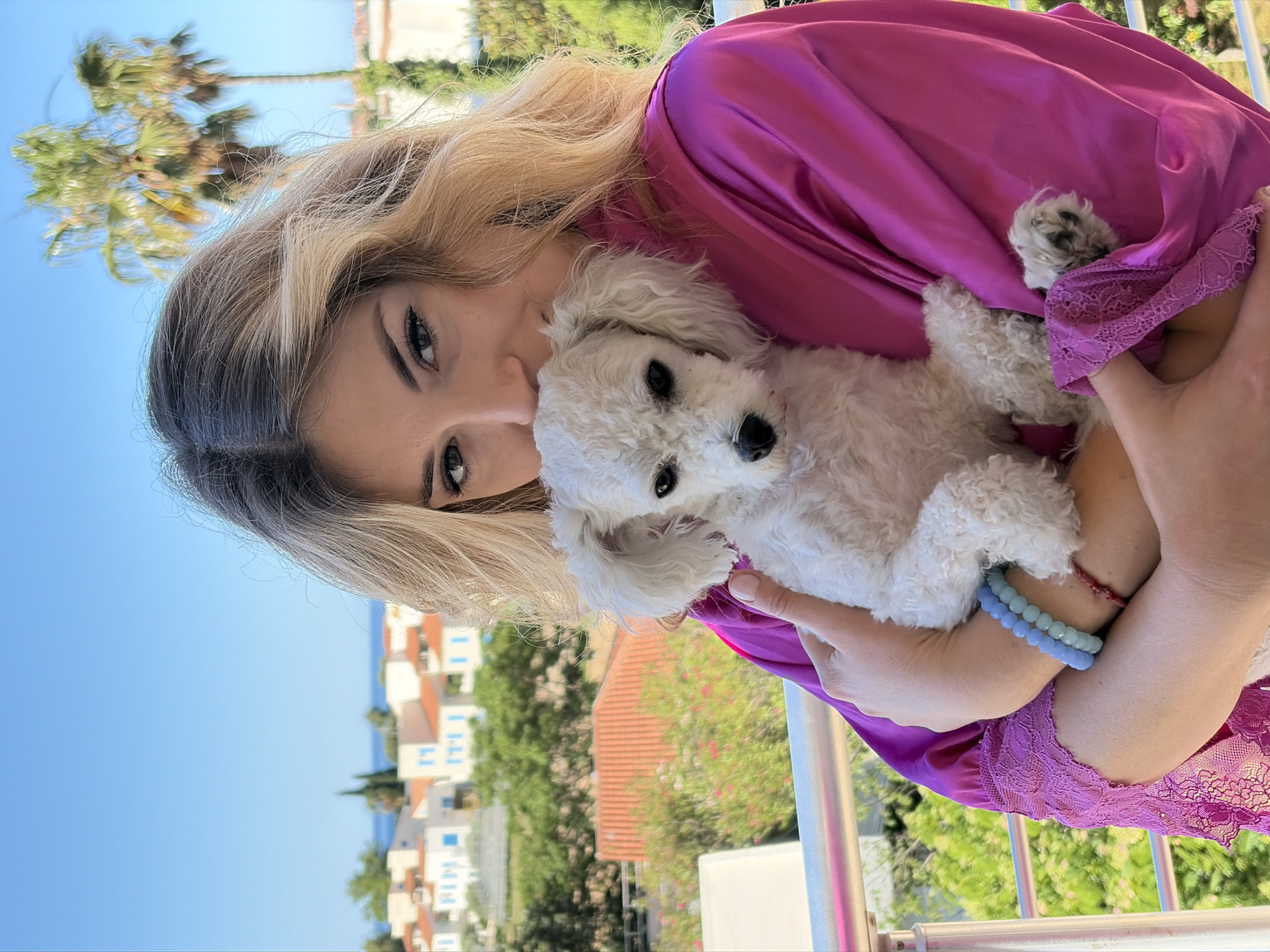 A woman wearing a fuchsia robe is holding a small, white poodle dog. She is outdoors on a balcony with a metal railing. In the background, there are white buildings with orange-tiled roofs, green trees, and a blue sky. The location appears to be Budva, Montenegro, based on the architectural style and coastal setting. The woman is looking at the camera. The poodle is also looking towards the camera.