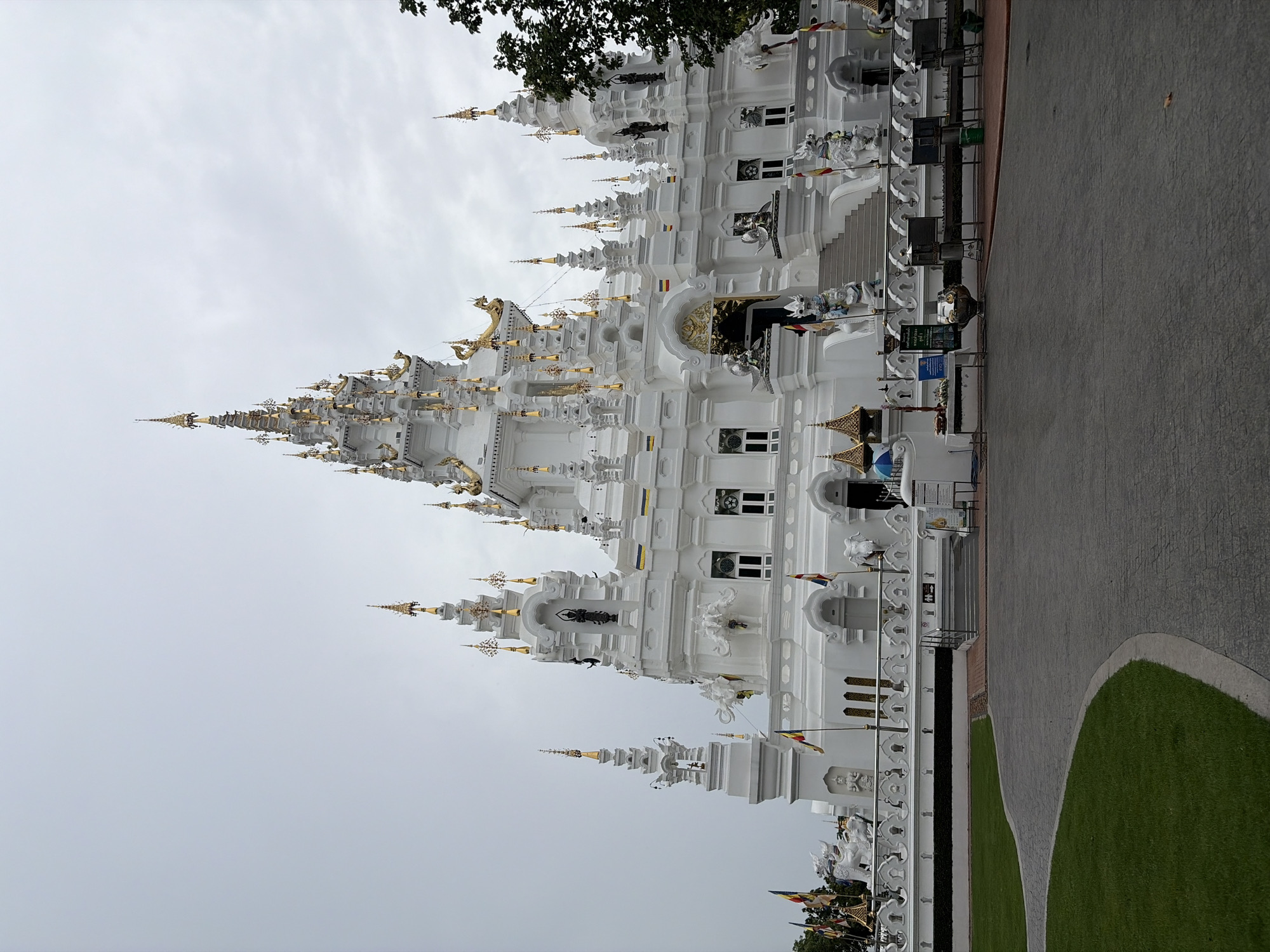 The image shows Wat Sriboonreuang, a white Buddhist temple in Ban Pong Nuea, Thailand, under a cloudy sky. The temple features ornate white structures with gold accents on its spires and rooflines. Decorative white elephant statues and other mythological creatures adorn the temple's exterior. The foreground includes a paved walkway and a manicured lawn. Various flags are displayed near the temple entrance, and a set of stairs leads up to a grand doorway. The scene appears to be devoid of people.