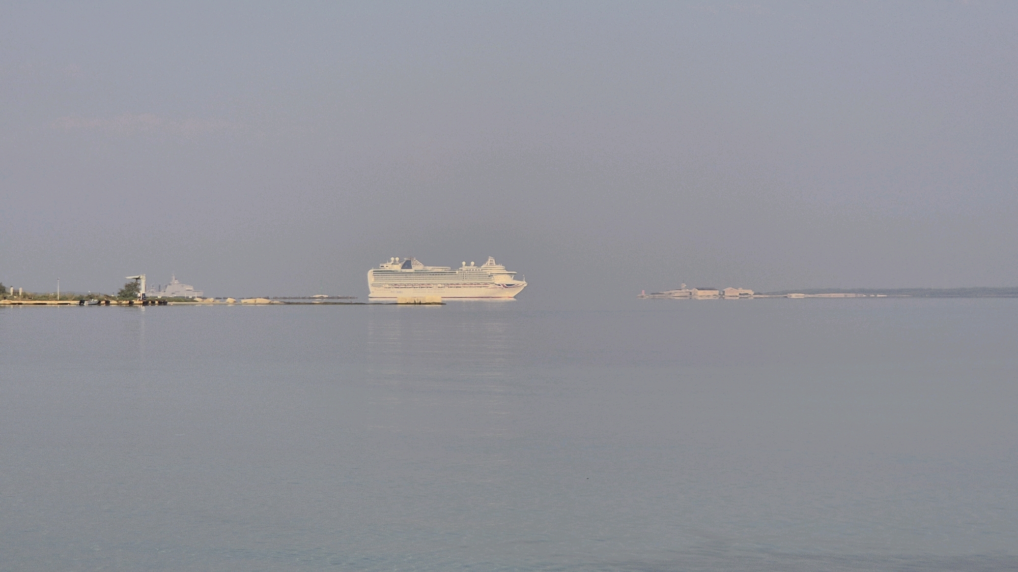 A wide, horizontal shot shows a calm body of water in the foreground with a faint, hazy sky in the background. In the mid-ground, a large, white cruise ship with multiple decks is the primary subject, sailing from left to right. To the left of the cruise ship, a gray naval vessel is visible near a small island with structures and vegetation. Further to the right, across the water, a long, low island with several buildings is discernible. The overall scene suggests a port or coastal area, potentially the Mar Piccolo or Mar Grande in Taranto, Italy, given the presence of both naval and civilian vessels, as well as industrial or port-like structures on the islands. The light is soft, indicating either early morning or late afternoon.