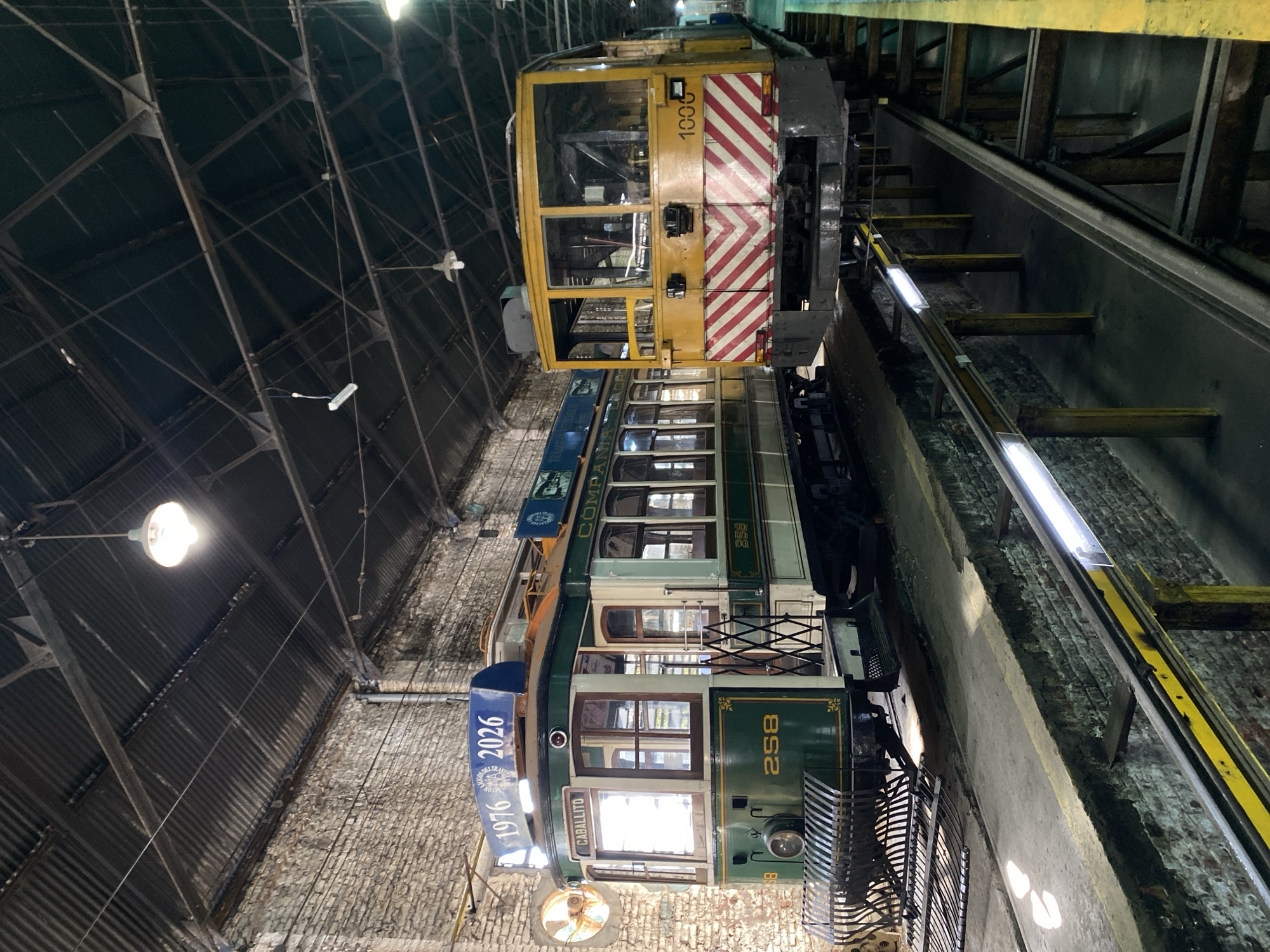 The image shows two tram cars inside a brick-walled structure with a high, arched metal ceiling. The lower tram car is green and labeled "258". A sign on the front of the tram reads "1976 2026." The upper tram car is yellow with red and white stripes and labeled "1000". These trams are historic and part of the Buenos Aires Tramway.