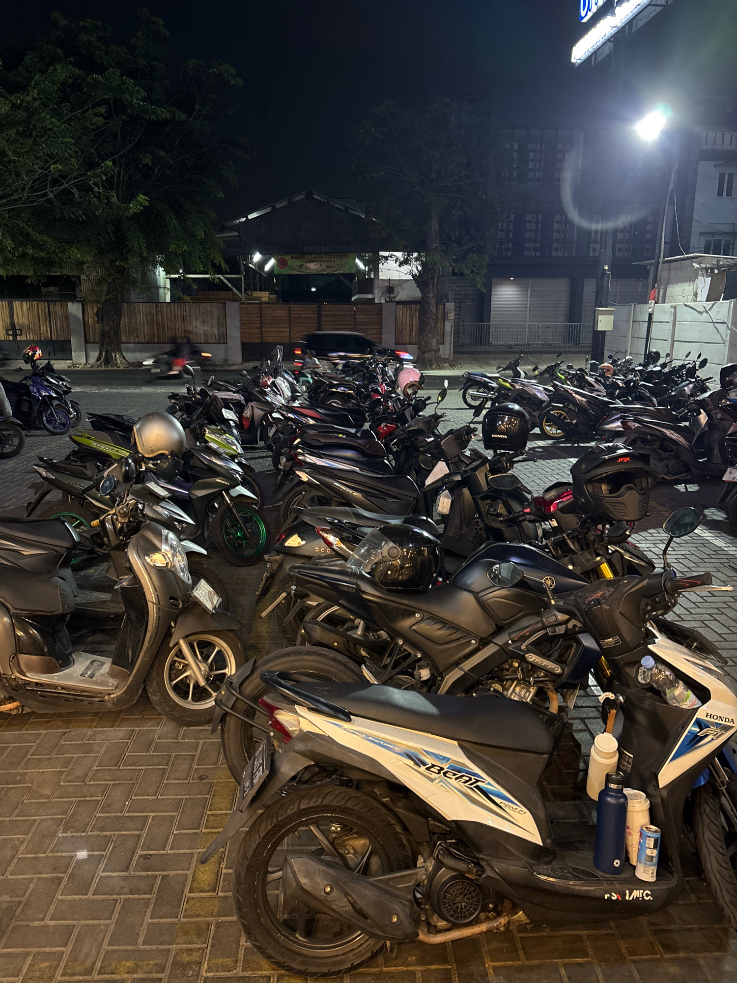 A wide-angle, low-light photograph captures a densely packed motorcycle parking lot at night, likely in Bekasi, Indonesia, indicated by the context. The foreground is dominated by several parked scooters and motorcycles, their bodies reflecting the ambient light. Several helmets are visible, some on seats and others on the ground. Two prominent water bottles and a can are placed on the floorboard of a white and blue scooter in the lower right portion of the frame. In the midground, rows of similar motorcycles are tightly arranged, with some individuals blurred due to motion, suggesting they are either arriving or departing. A few trees with dark foliage line the perimeter of the parking area. In the background, a building with a dark facade and illuminated windows is visible, along with a streetlamp casting a bright, diffused light upwards. The ground is paved with interlocking brick pavers.