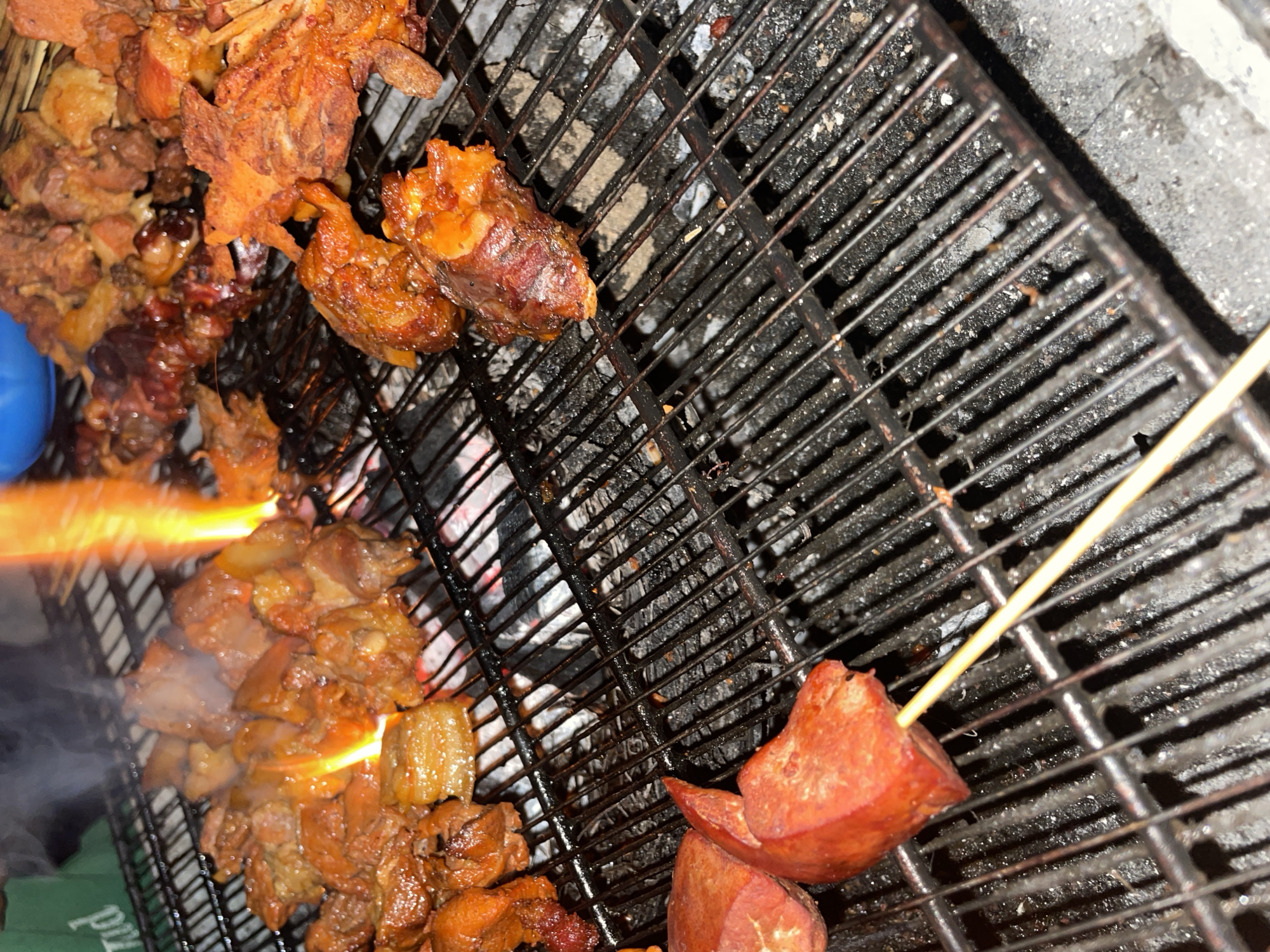 The image depicts food being grilled on a charcoal grill. Visible are various cuts of meat and sausage being cooked. Flames are present, indicating the grilling process. The image is captured at an angle, showing the grill grates, food, and surrounding area. The image appears to be taken on Rukuba Road, Nigeria.