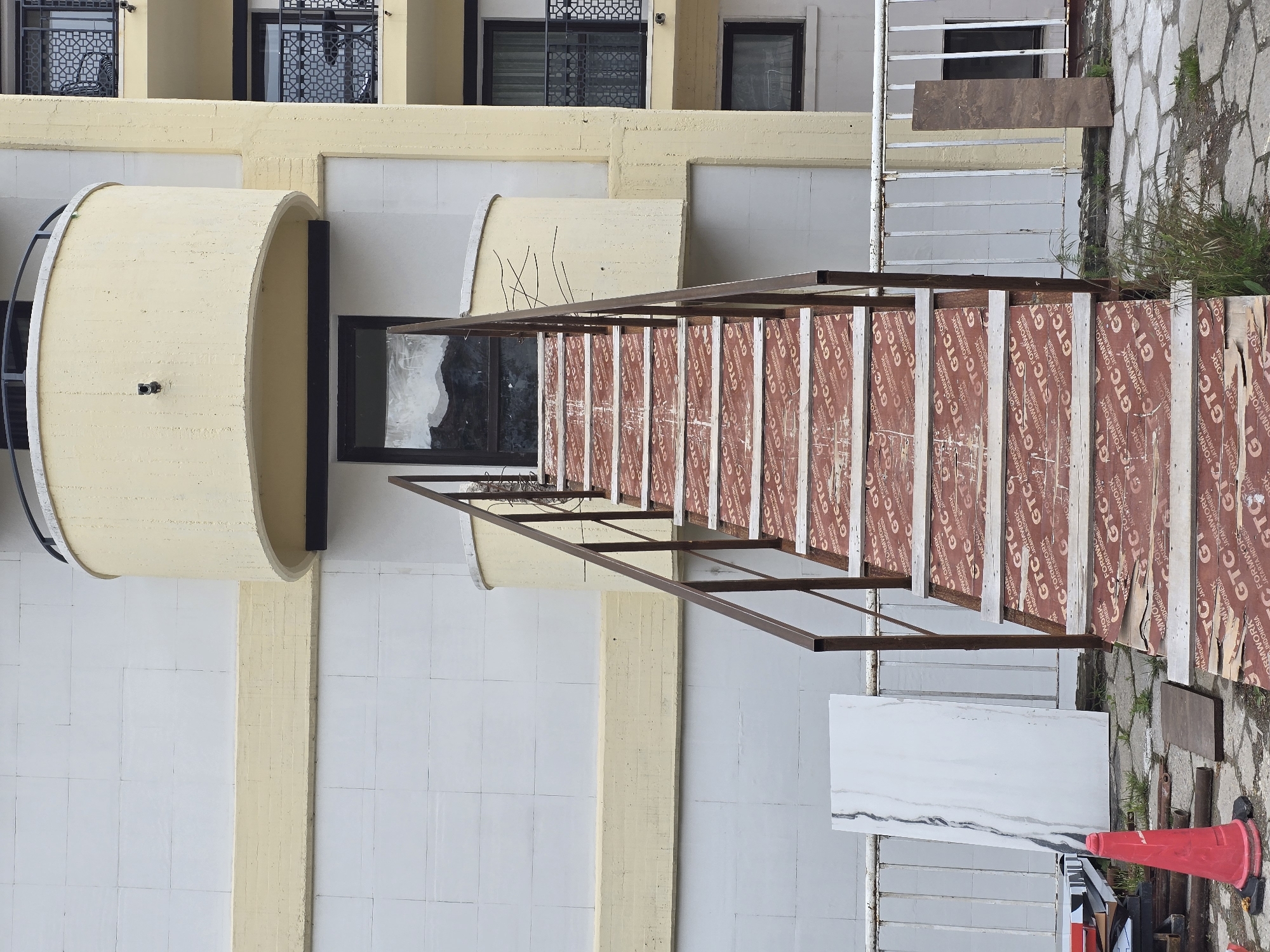 Here's a matter-of-fact description of the image:
The image depicts the exterior of a building under construction, likely in an urban setting. The building has a light-colored facade, featuring both smooth and textured surfaces. A cylindrical structure projects from the building, with a window nearby. A makeshift scaffold, constructed with metal frames and plywood sheets, extends from the building towards the foreground. The plywood is printed with the text "GTC FORMWORK," suggesting its use in concrete forming. The scaffold appears to be set up on a paved area, with construction materials, including a red traffic cone and a marble slab, visible nearby. The overall scene indicates an active construction site, potentially in the process of renovation or expansion.